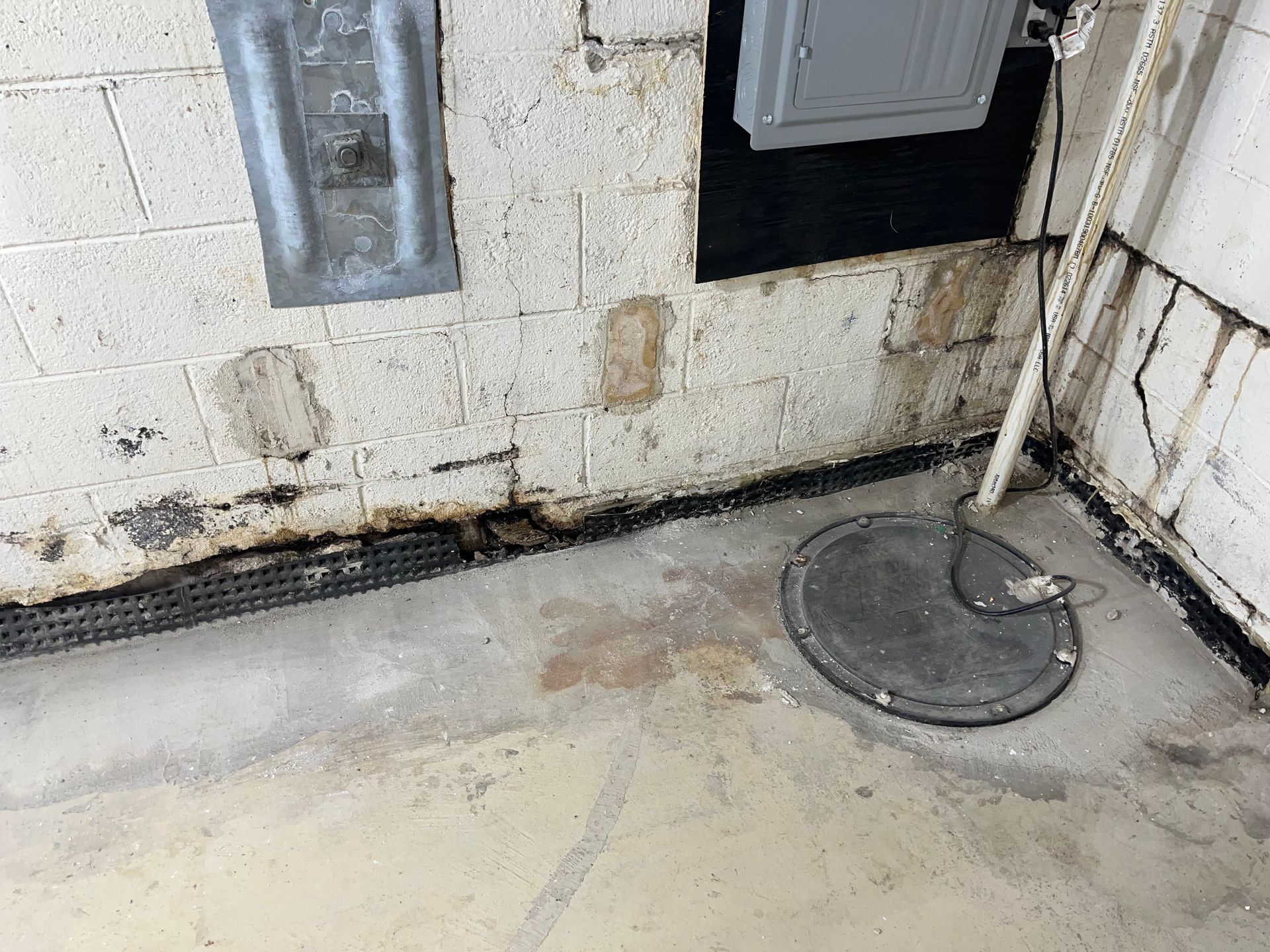 Basement utility room with stained cinderblock wall, electrical panels, and a floor drain cover.