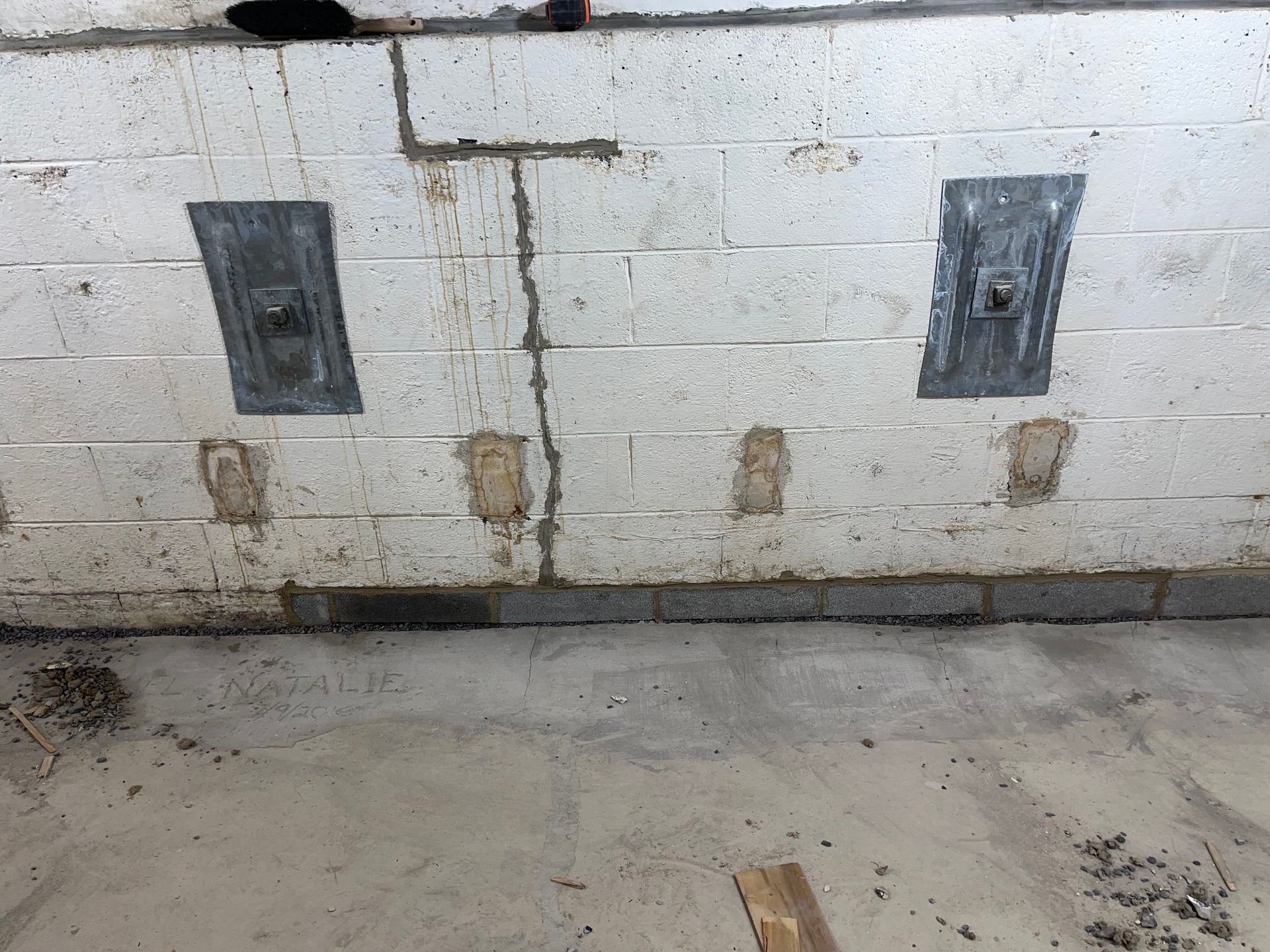 Basement cinder block wall with two metal plates, a floor drain, and water stains on concrete floor