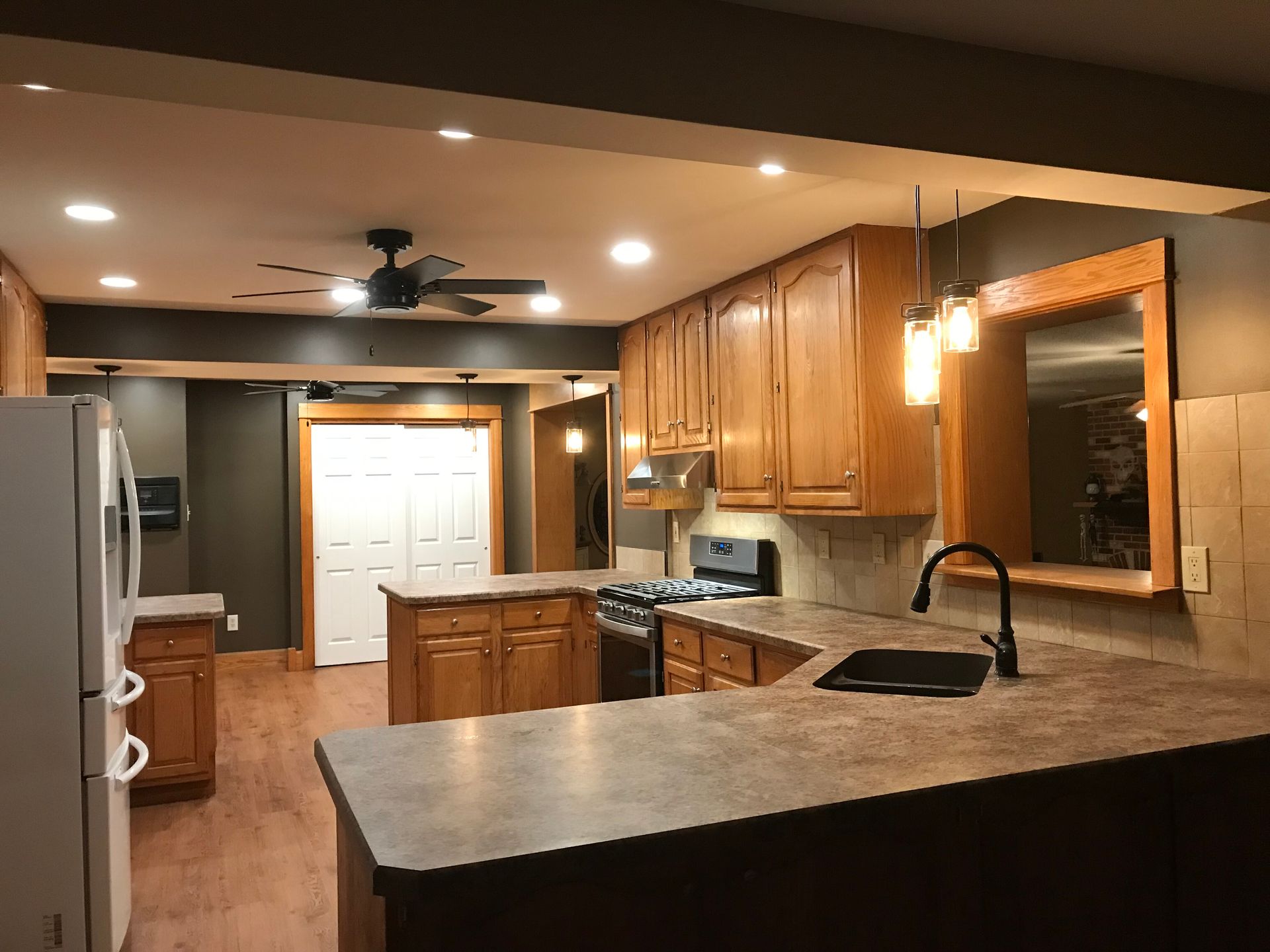 Modern kitchen with wood cabinets, granite island, and recessed lighting, viewed toward a white door and stainless fridge