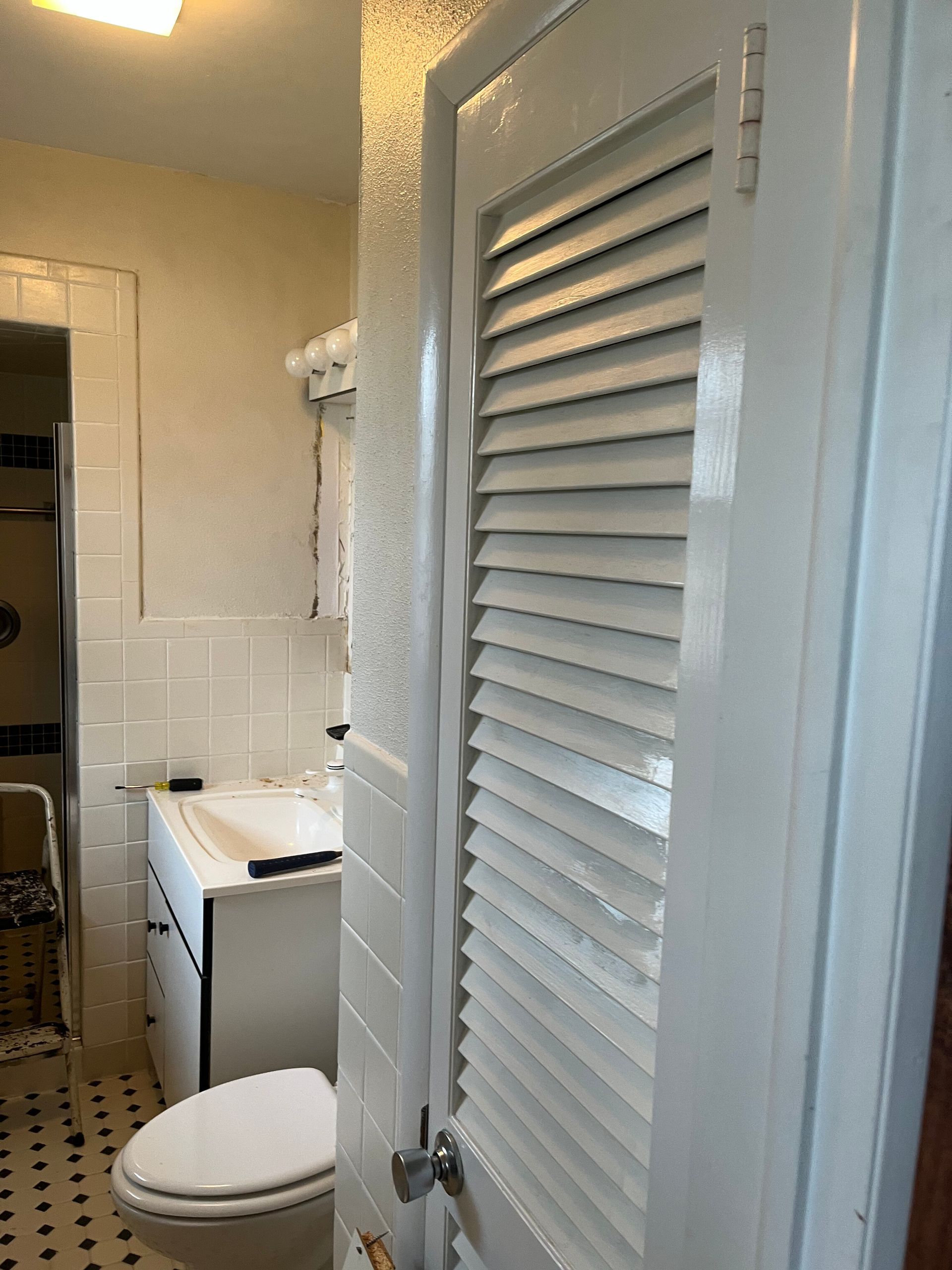 Small bathroom with toilet, sink, tiled shower, and white louvered door in the foreground