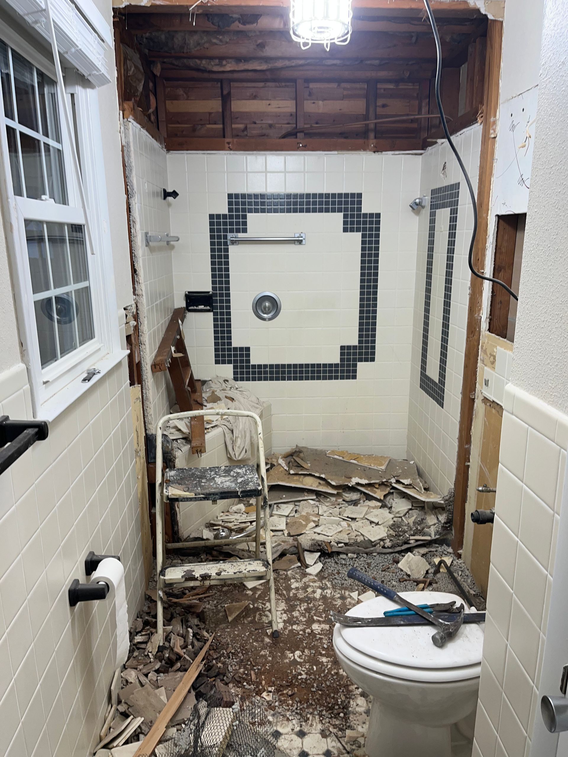Narrow bathroom under demolition with broken tile, exposed ceiling, and a toilet in the foreground