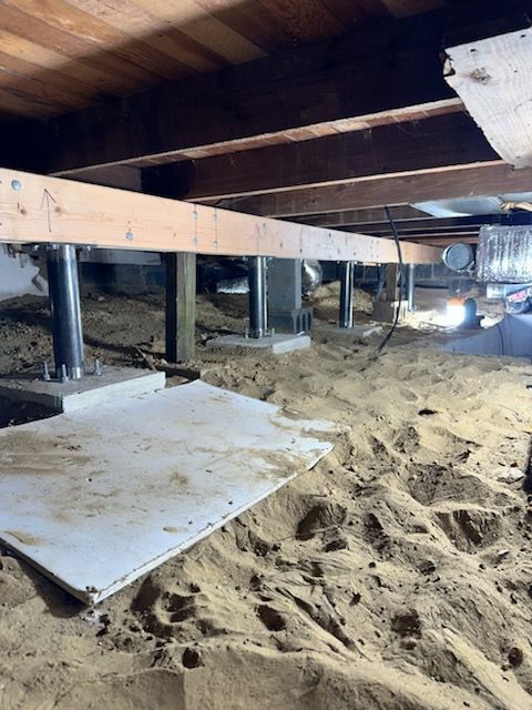 Under-house crawlspace with wooden beams, support posts, insulation, and sandy ground cover.