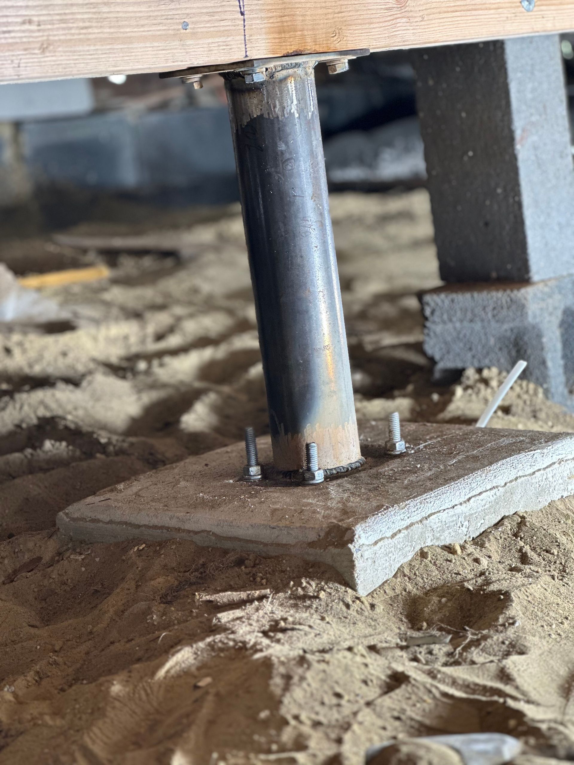 Metal support post under a raised wooden floor on sandy ground, with bolts and concrete footing.
