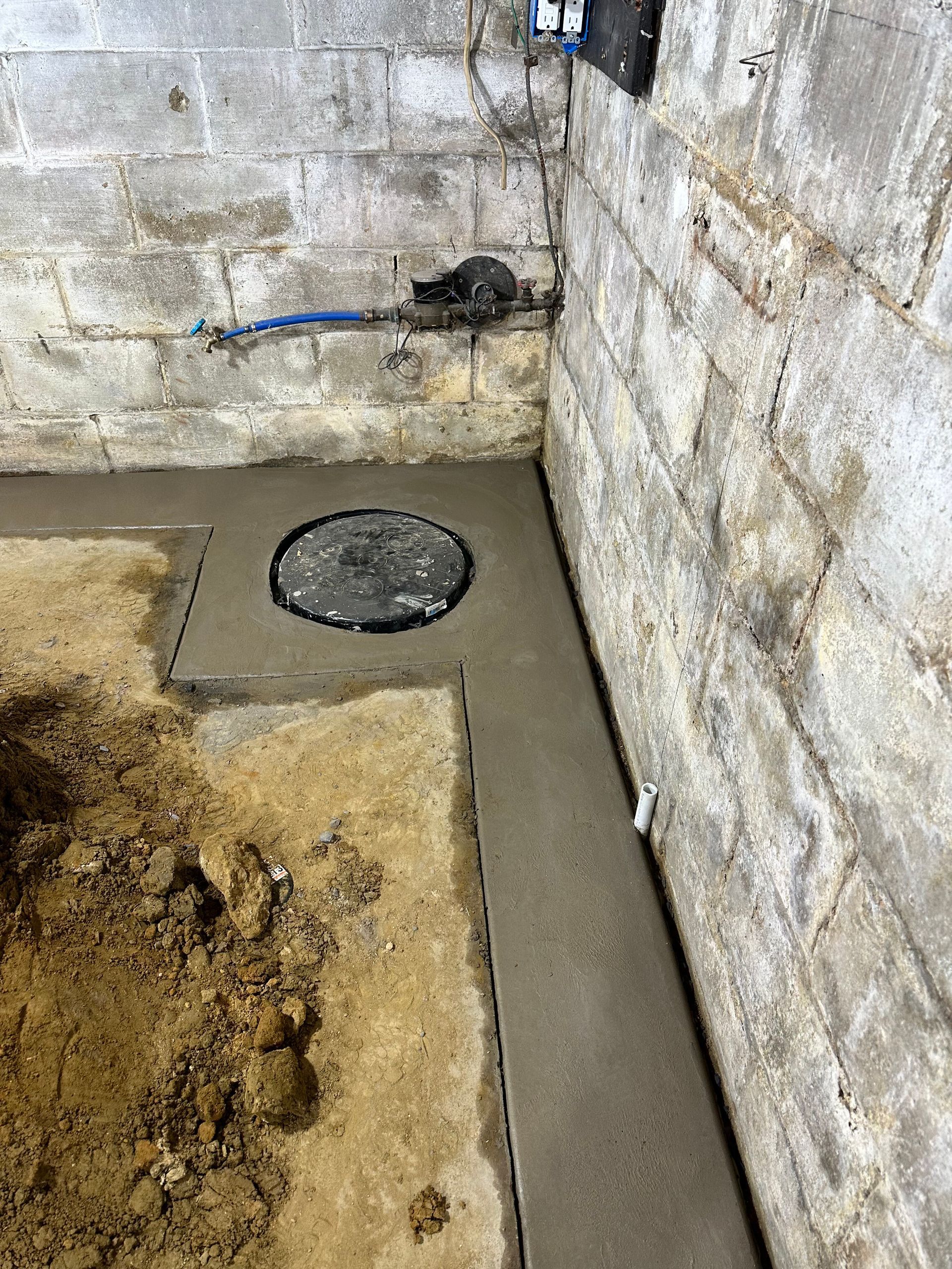 Concrete basement corner with a round floor drain and exposed blue pipe on rough cinderblock walls