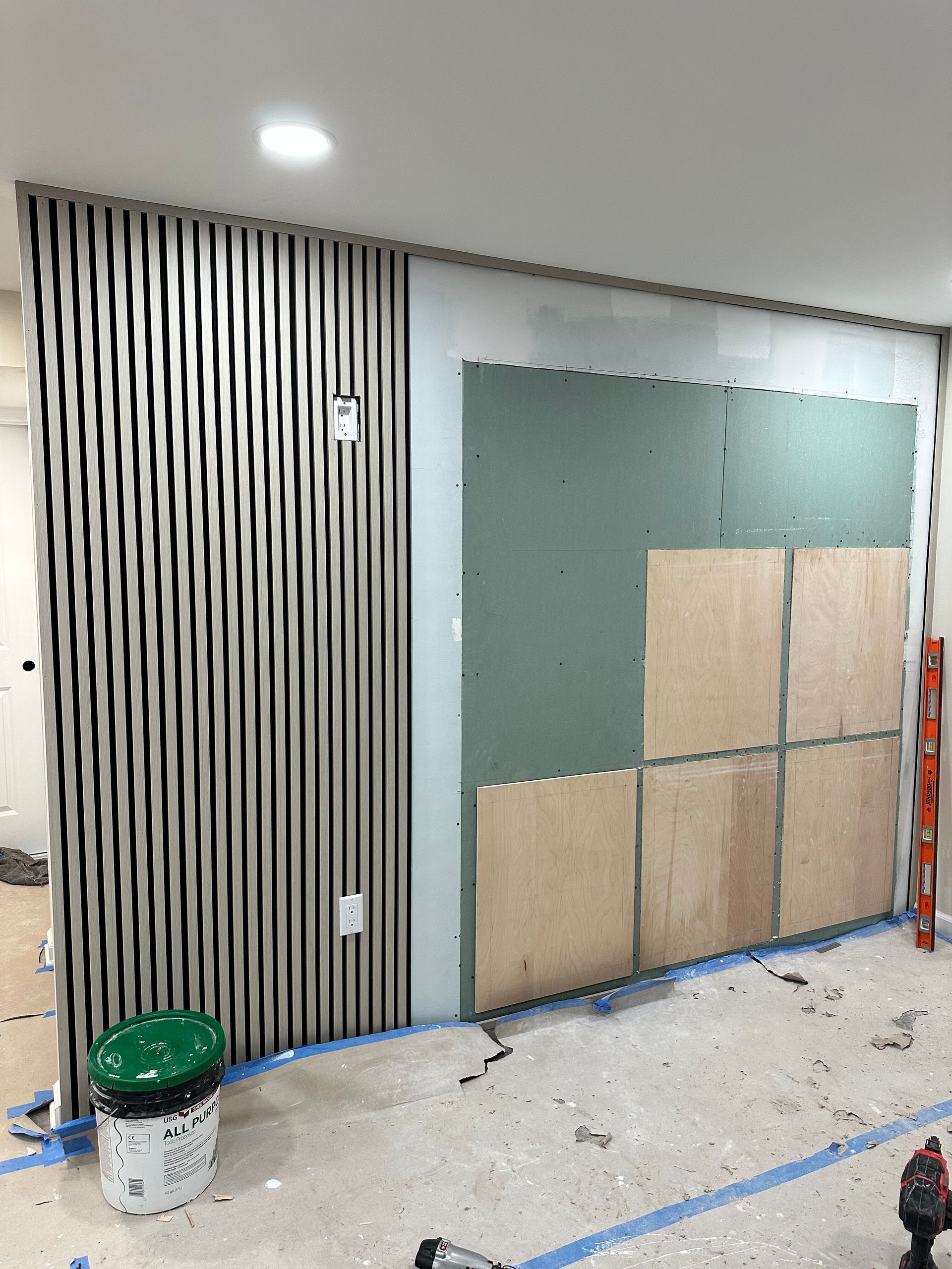 Partially renovated room with striped wall paneling, green board, protective floor covering, and paint bucket