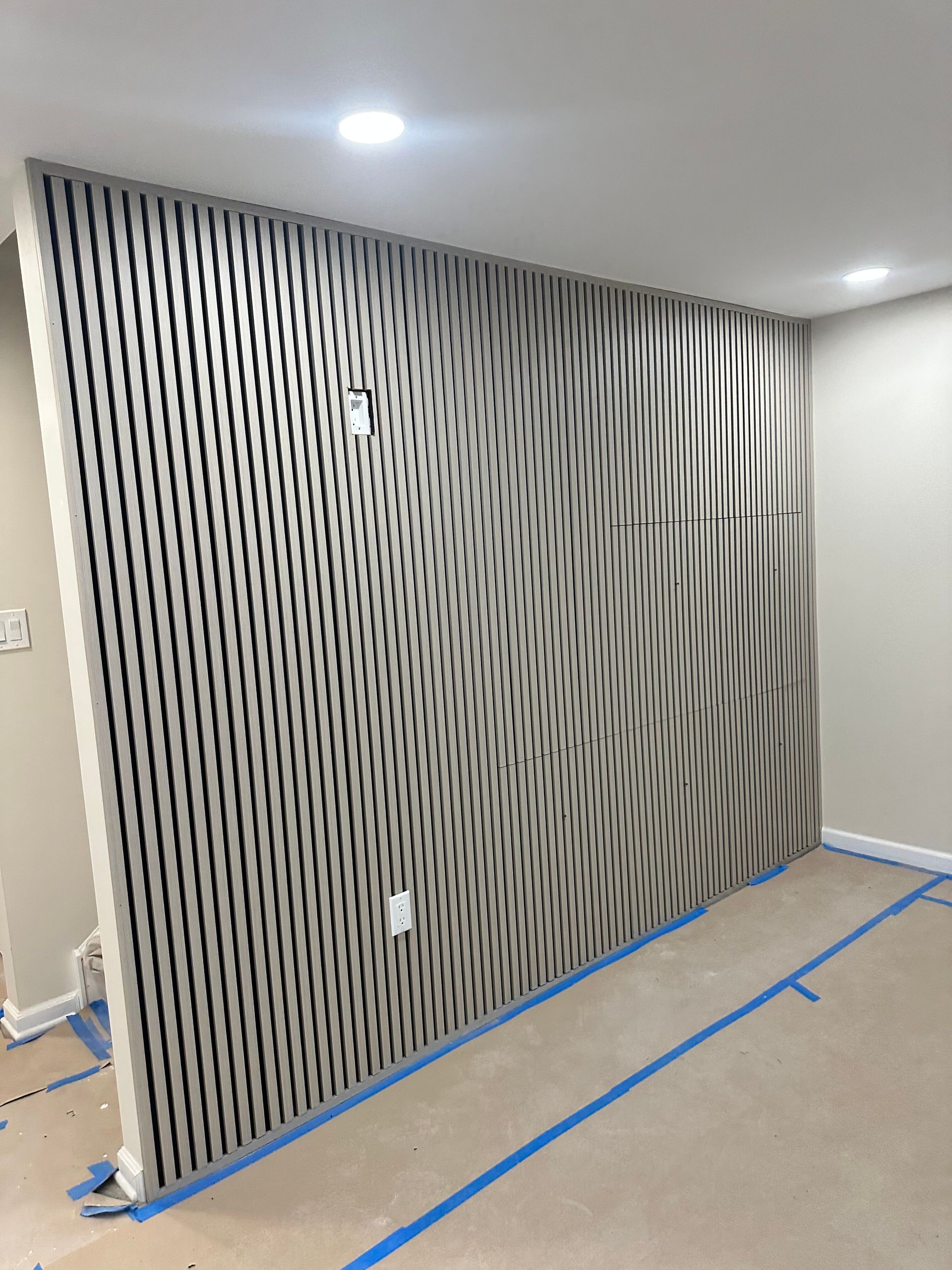 Decorative gray slatted wall panel in a room, with blue tape on the floor nearby
