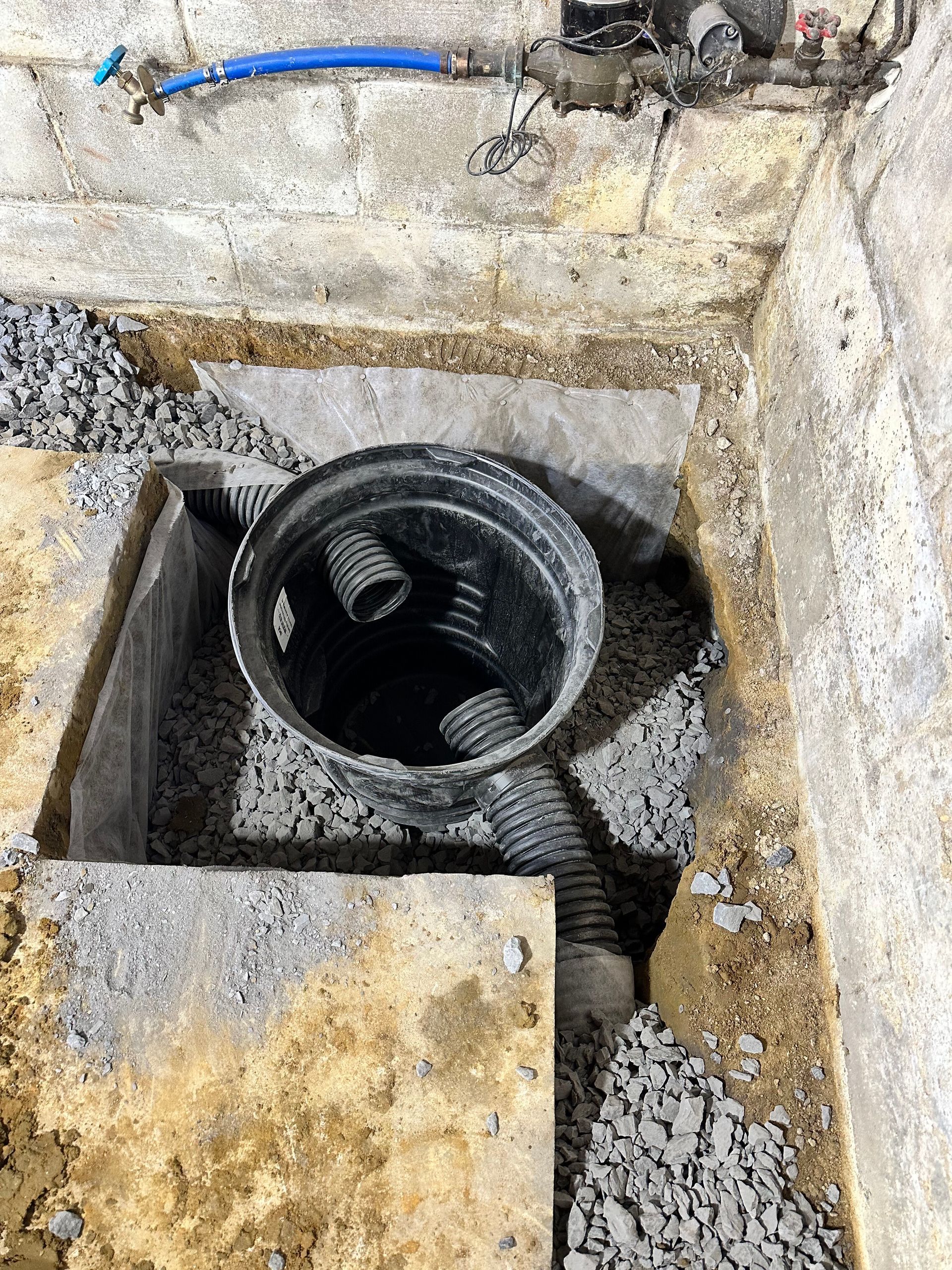 Open utility hole with black pipe and corrugated hose in a rough concrete floor area