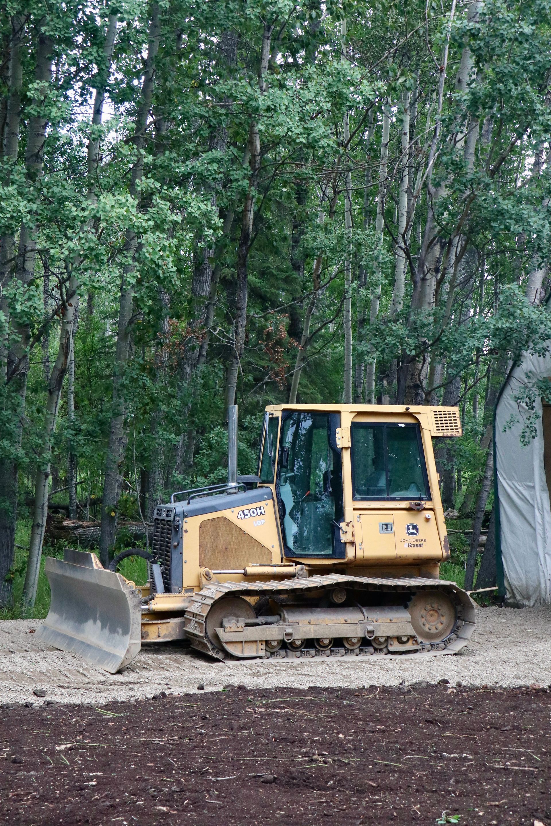 Yellow bulldozer on a dirt lot beside a wooded area, with a raised blade and forest backdrop.