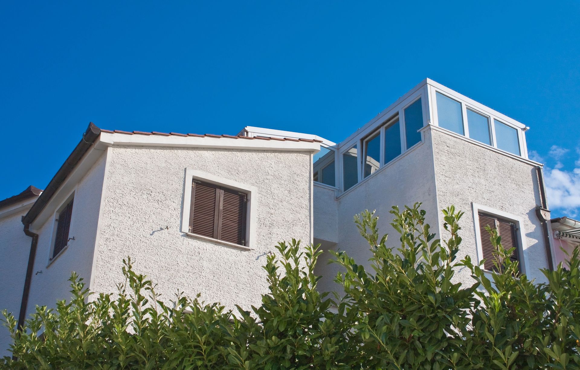 Casa in stucco bianco con persiane marroni e tetto vetrato, sullo sfondo di un cielo azzurro brillante.