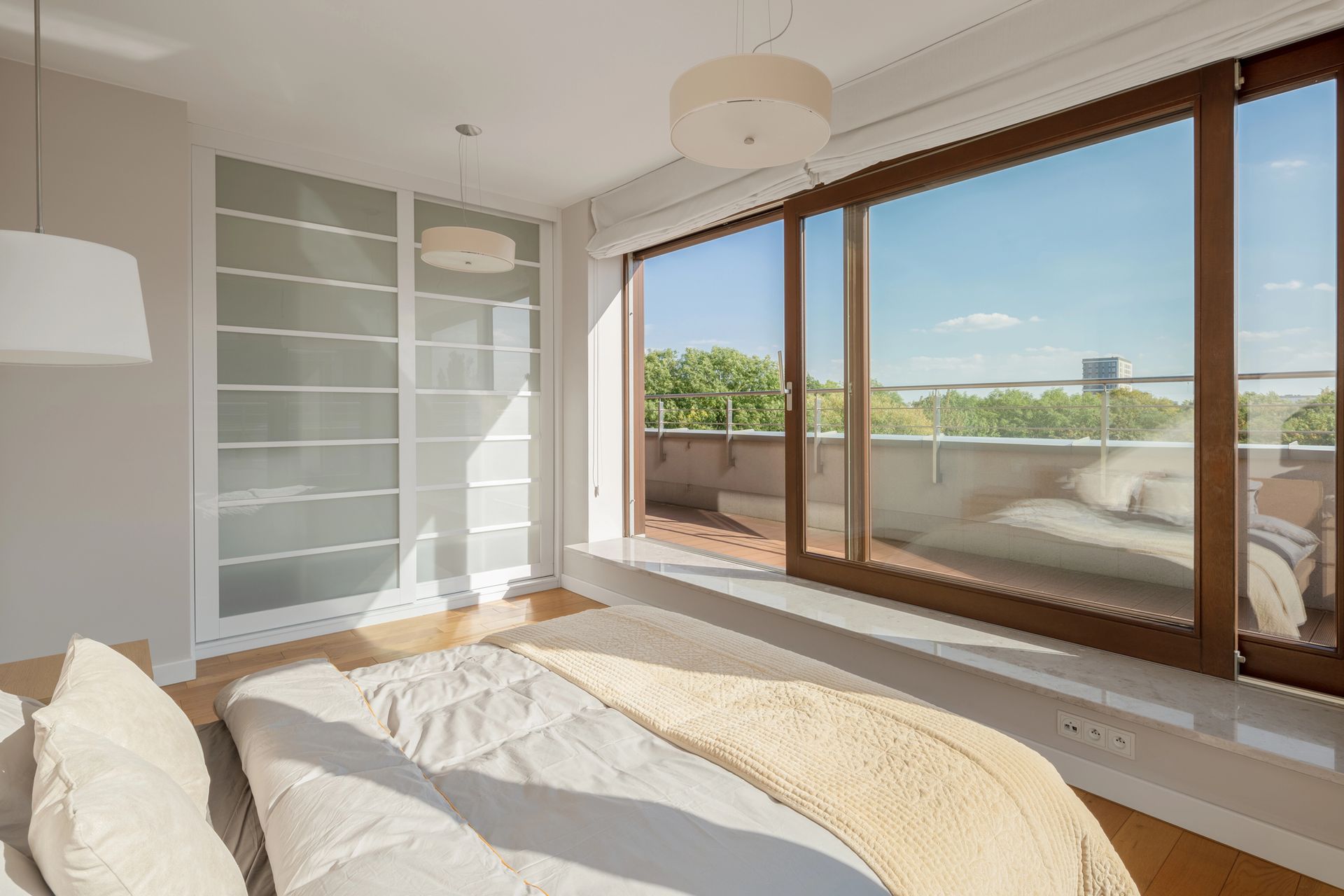 Camera da letto luminosa con ampie finestre, letto bianco e balcone con vista soleggiata sulla città o sul mare.