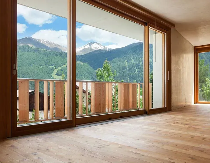 Luminosa stanza vuota con ampie finestre con infissi in legno che si affacciano su verdi montagne e un balcone.