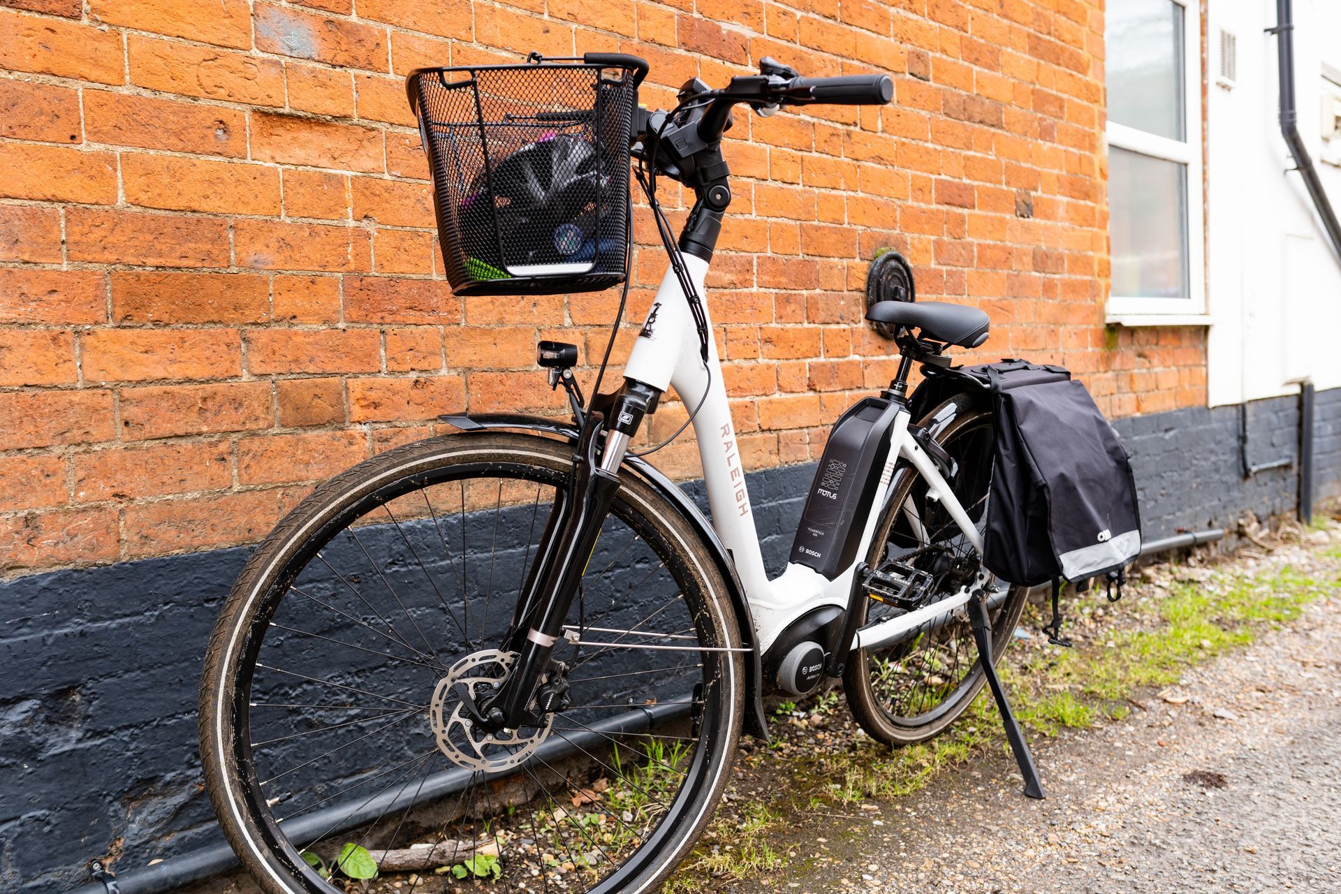 White city bike with front basket and rear panniers leaning against a brick wall outdoors