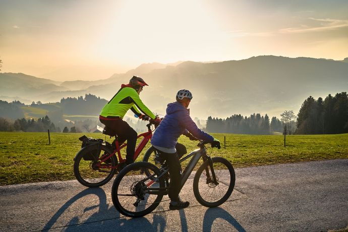 Two cyclists ride along a rural road at sunrise with mountains in the background.