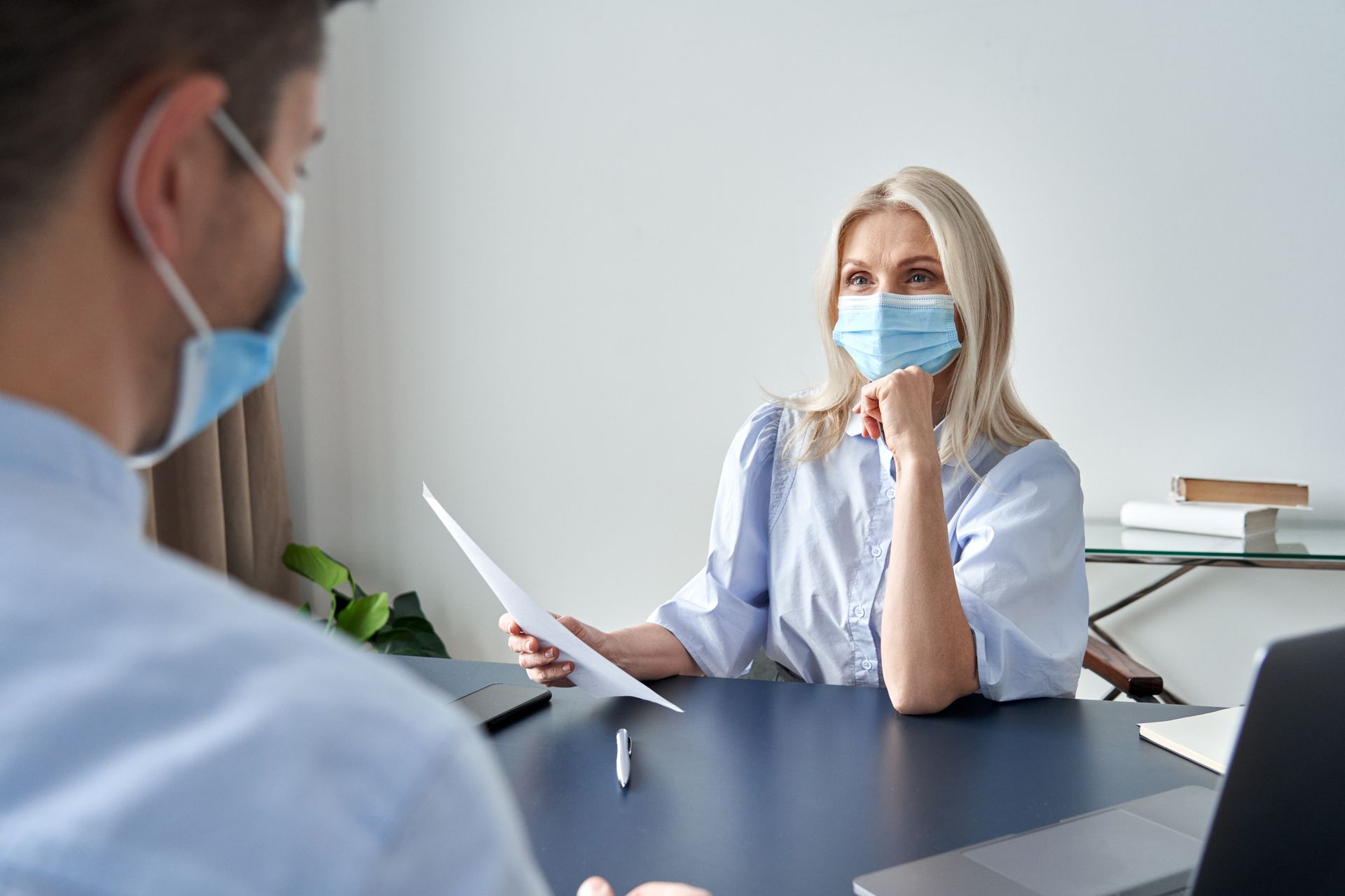 Female hr specialist manager, employer wearing face mask holding cv listening male candidate at job interview meeting