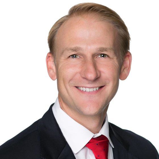 Smiling person seated against a light gray background, wearing a light blue button-up shirt and dark pants