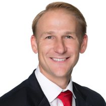 Headshot of a smiling man in a dark suit, white shirt, and red tie on a plain background