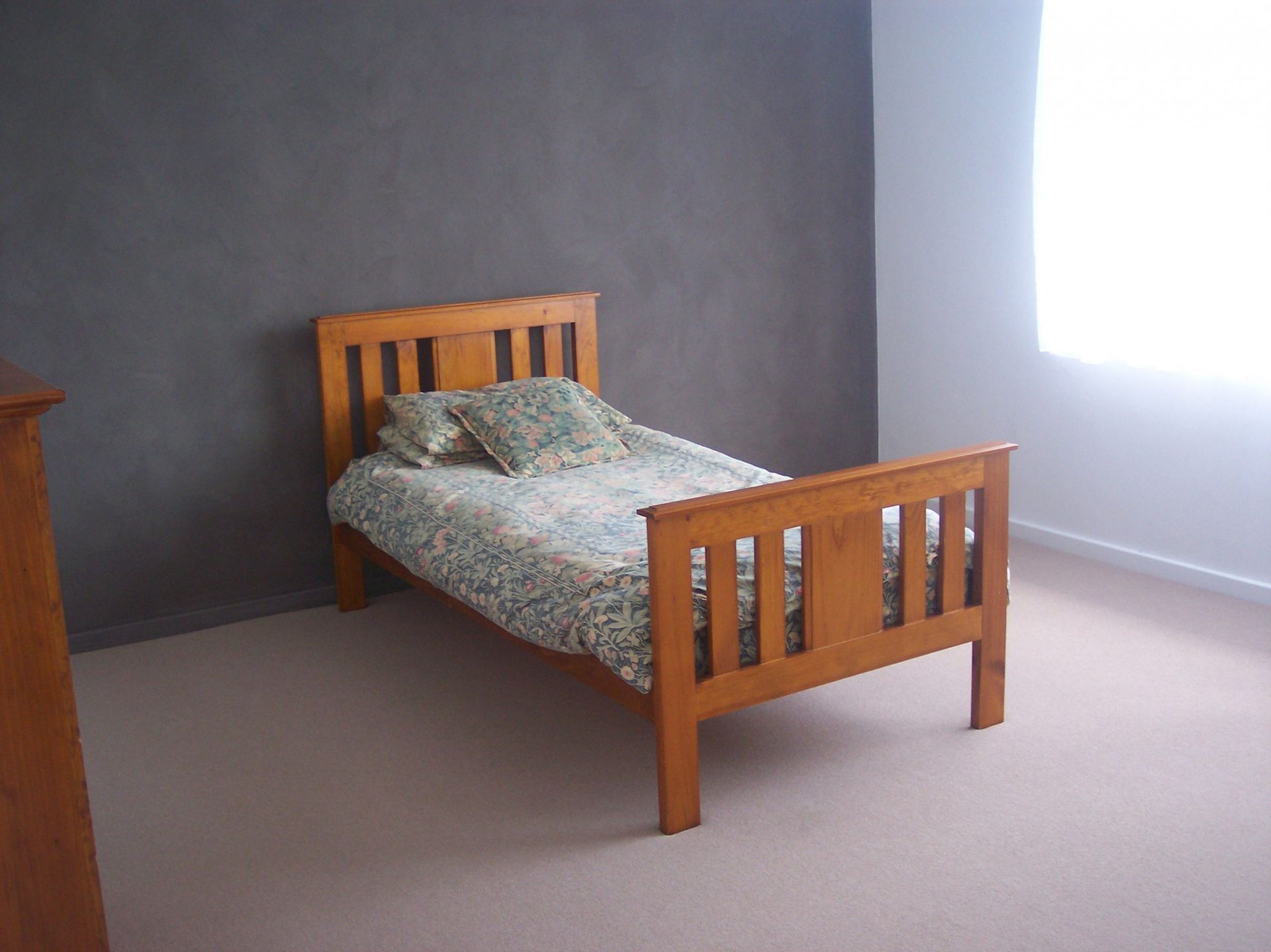 Wooden bed with patterned bedding in a sparsely furnished room with gray walls and a bright window