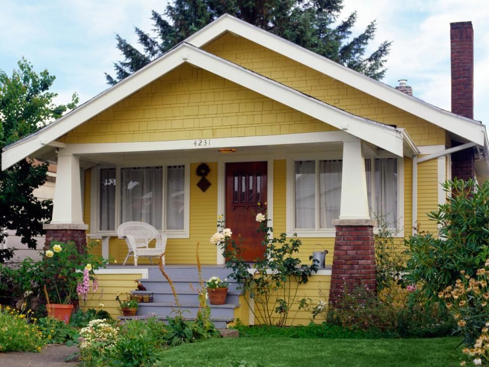 Yellow bungalow with red door, white trim, and a small front porch surrounded by lush garden greenery
