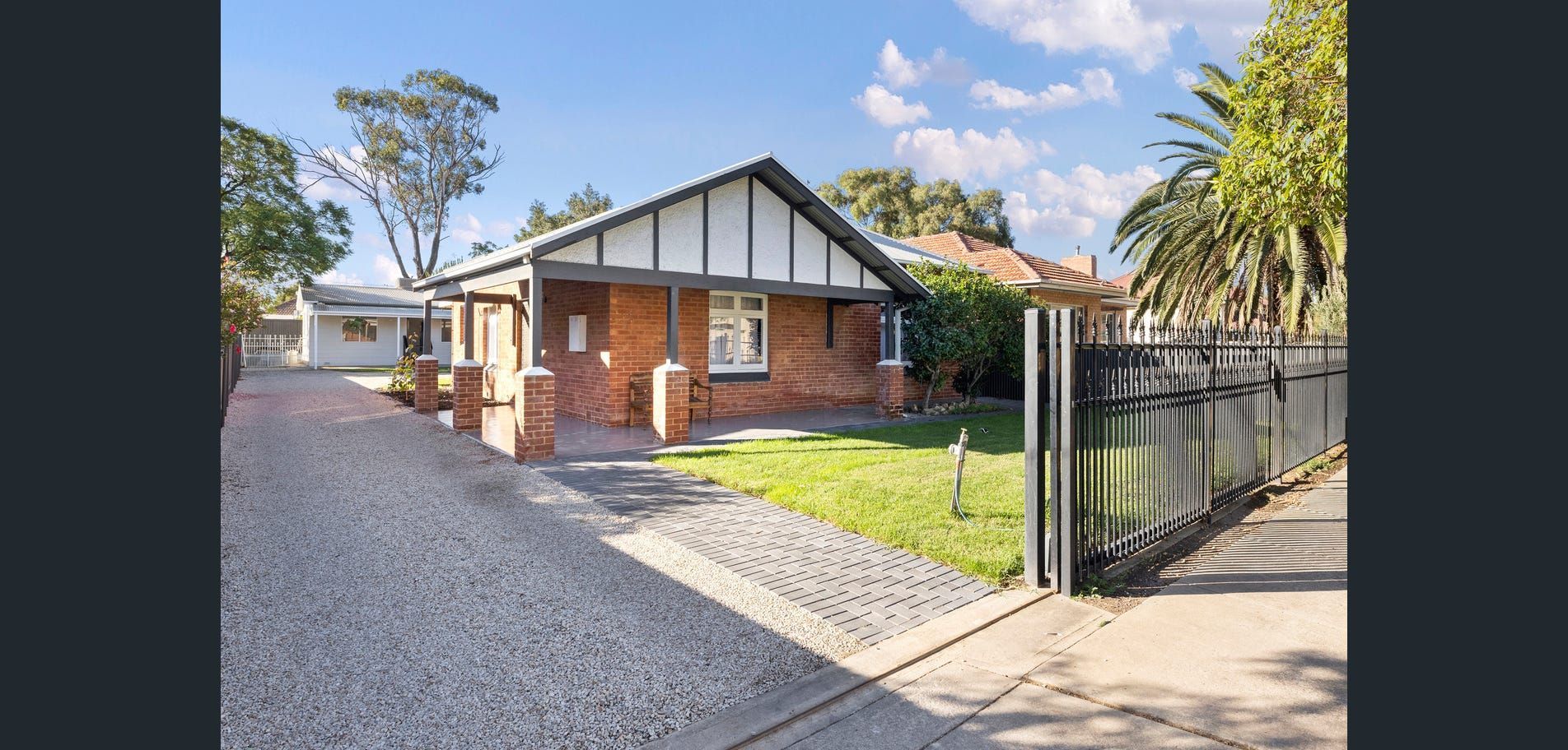 Brick house with white gable trim, front lawn, driveway, and open gate on a sunny day