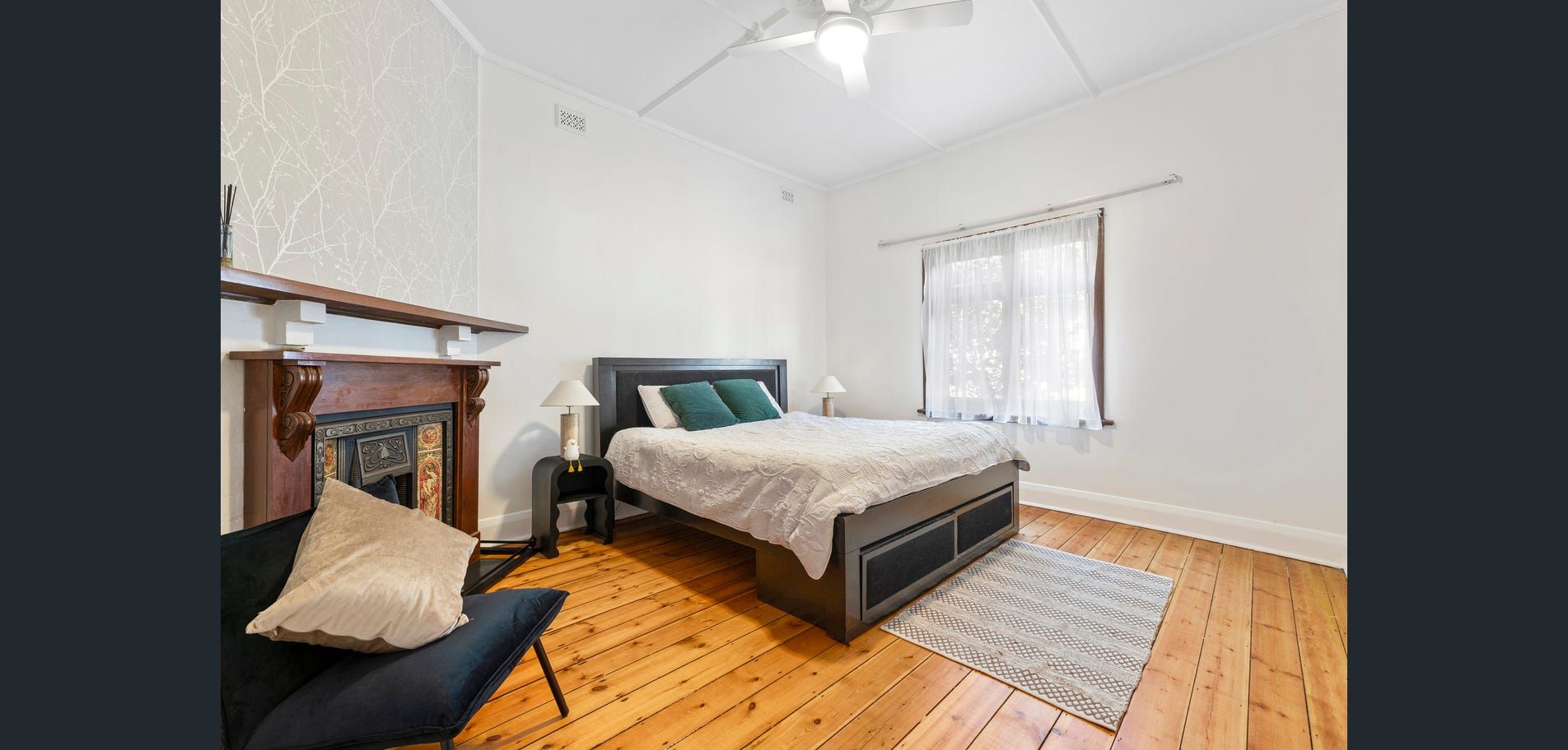 Bright bedroom with bed, armchair, fireplace, hardwood floors, and white walls, lit by a window