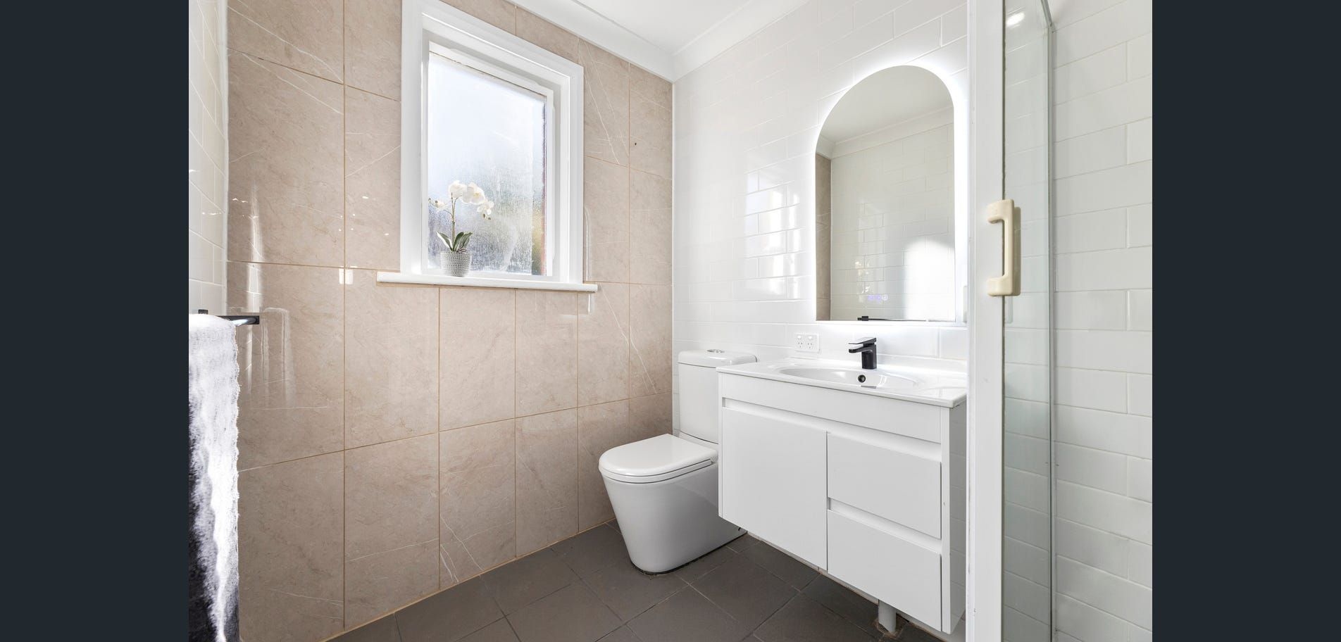 Bright beige bathroom with toilet, white vanity, arched mirror, and window under a sloped ceiling