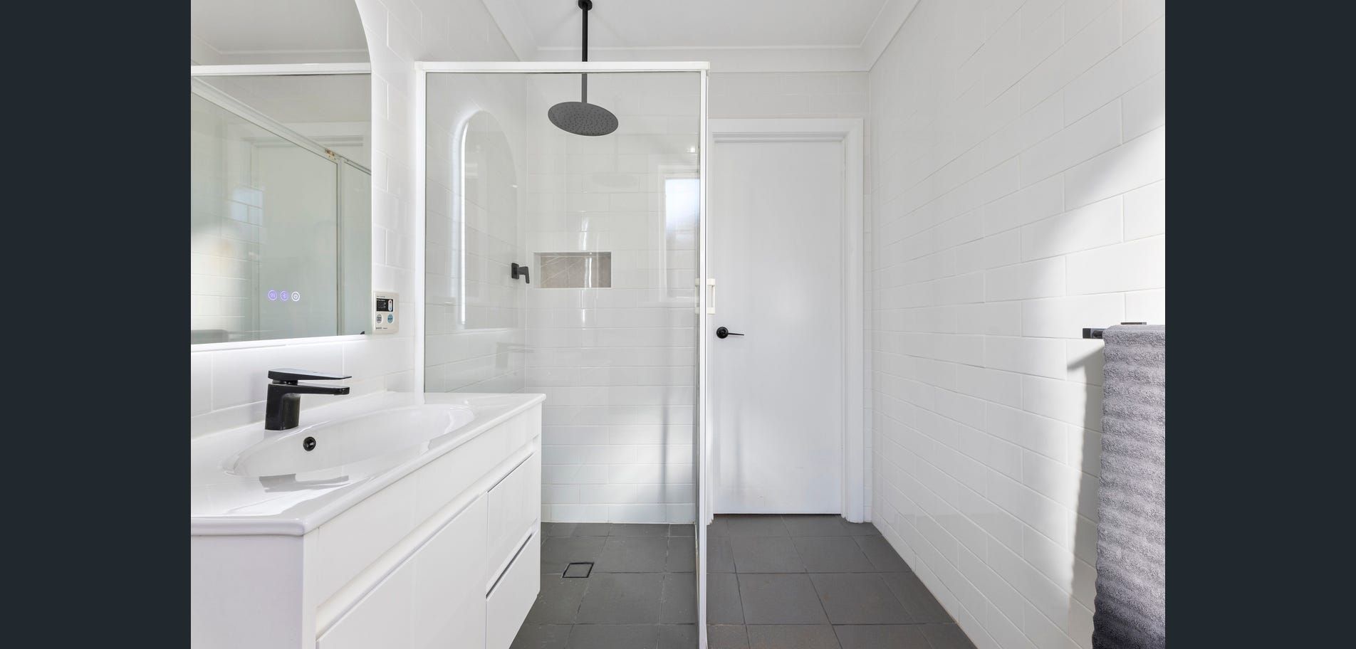 Bright white modern bathroom with double vanity, glass shower, and dark floor tiles.