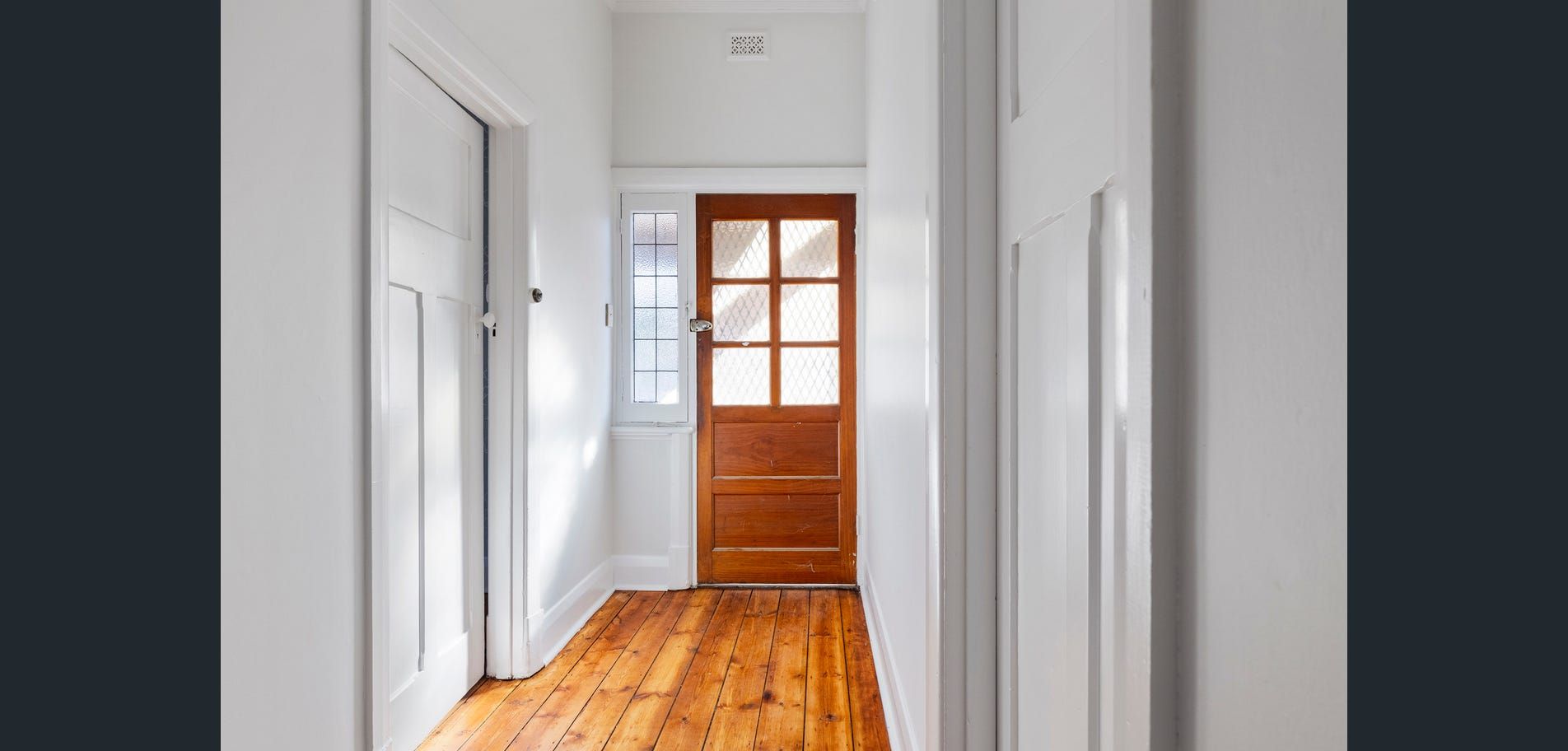 Bright hallway with white walls, wooden floor, and a wooden front door at the end.