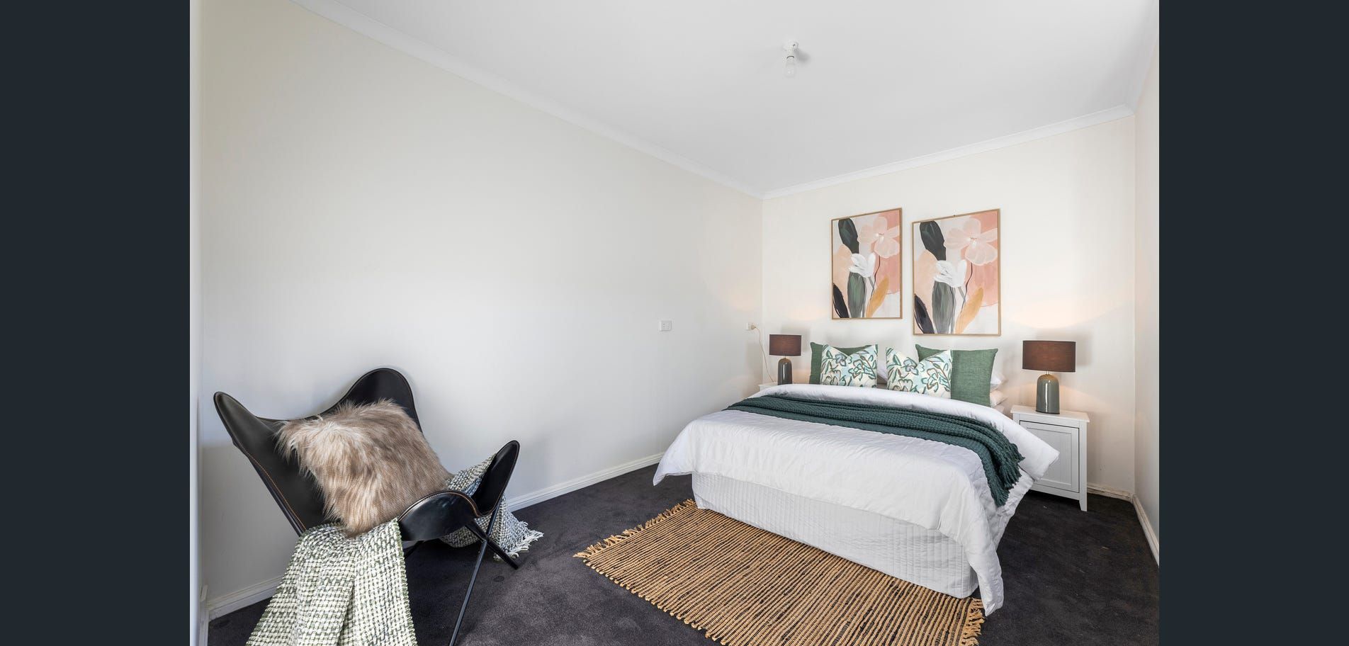 Minimal bedroom with white walls, black bed, abstract wall art, wicker rug, and a black chair with a cushion