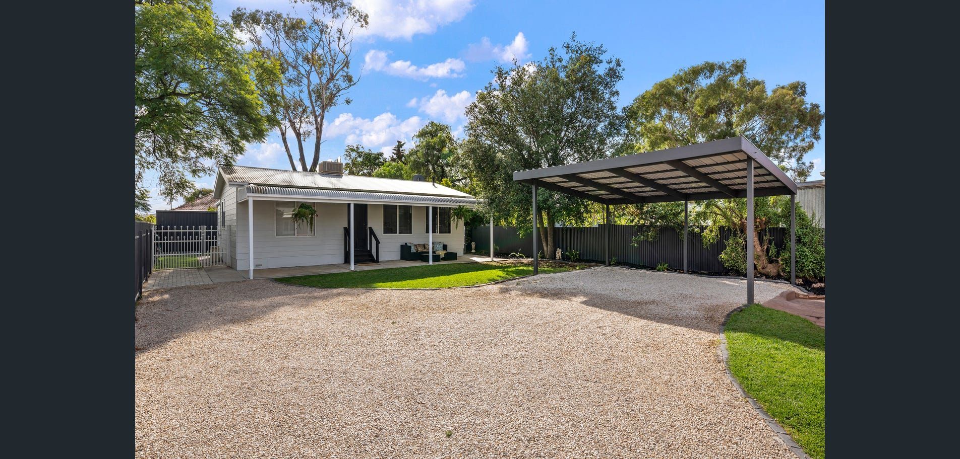Single-story house with gravel driveway, carport, and trees under a blue sky