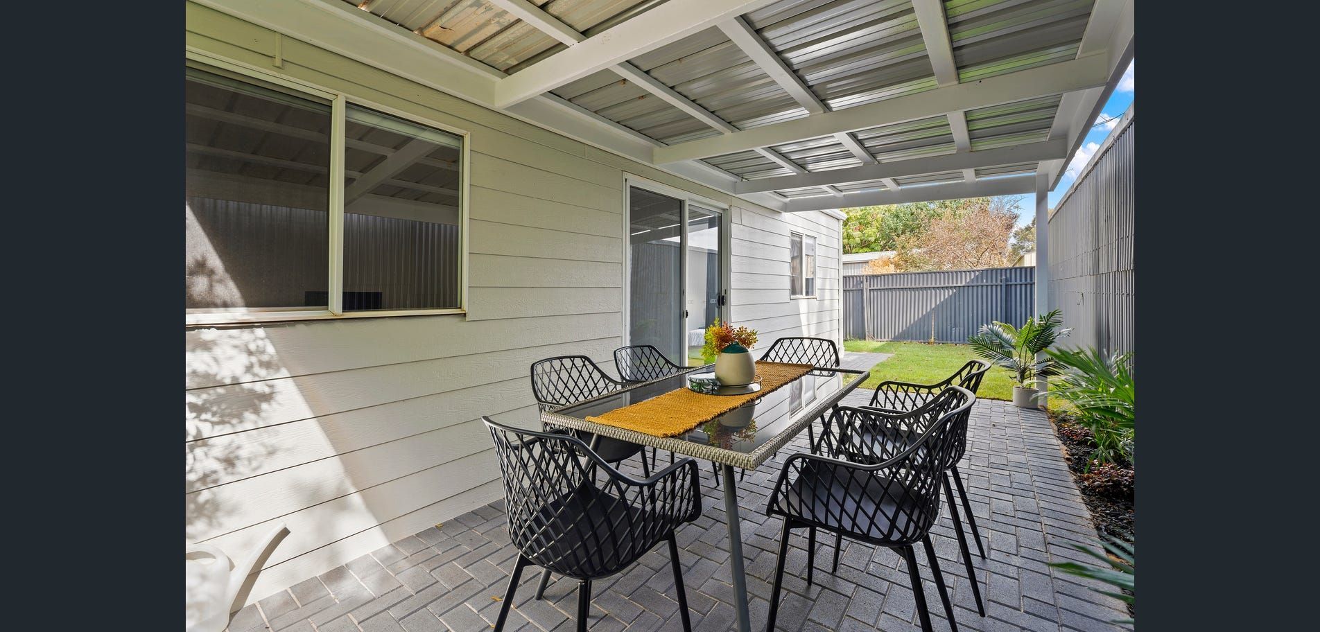 Covered patio with black chairs, wooden table, and a yellow vase beside a house wall and sliding door