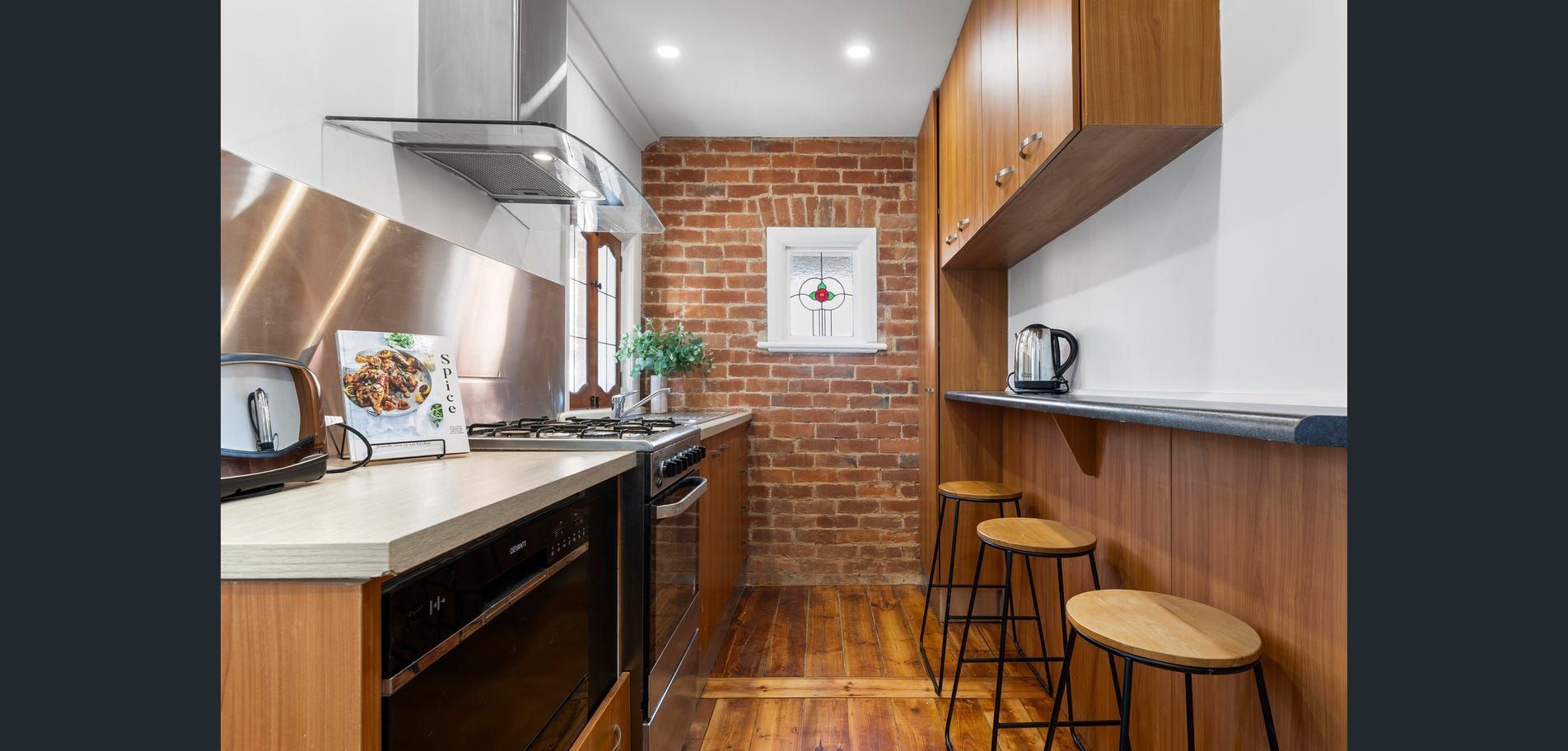 Narrow modern kitchen with exposed brick wall, wood counters, stainless appliances, and two stools.