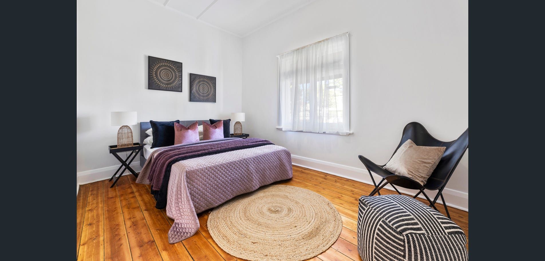 Bright bedroom with a bed, patterned rug, desk, chair, and large window on hardwood floors