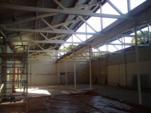 Abandoned warehouse interior with exposed roof trusses, scaffolding, and sunlight on the floor