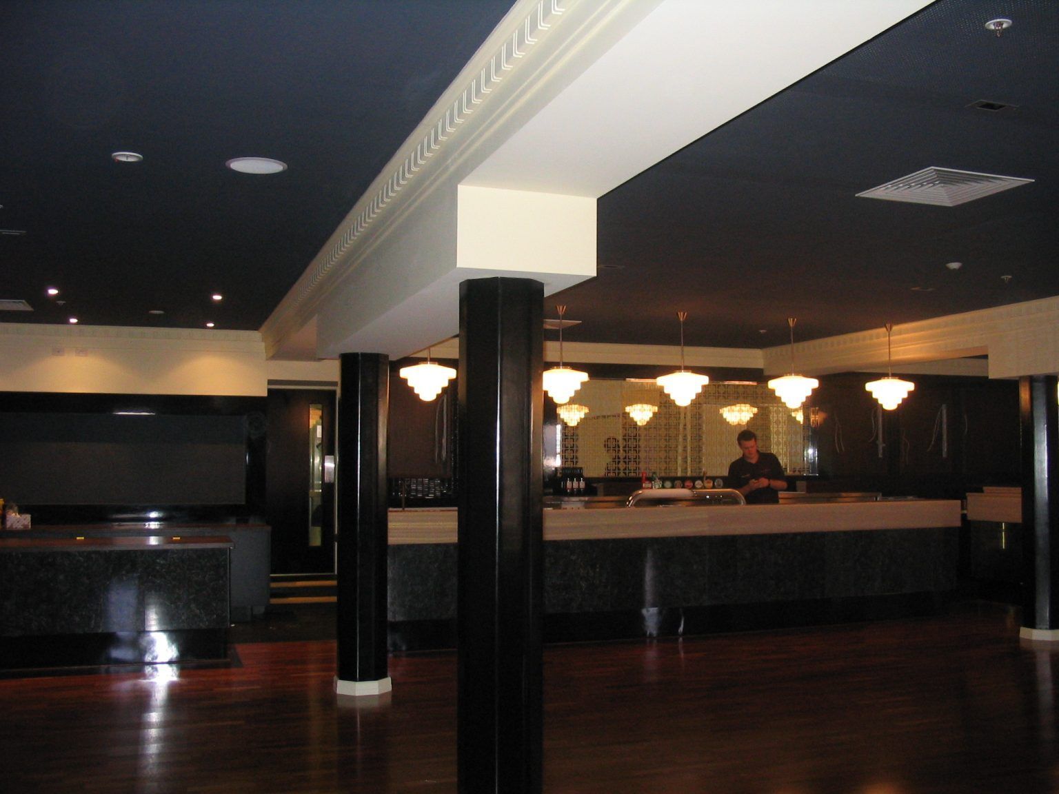 Dim hotel lobby with dark wood floors, black columns, and a reception desk under warm lights.