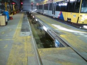 Train maintenance pit beside a yellow tram in a depot, with a concrete platform and rails running through the center
