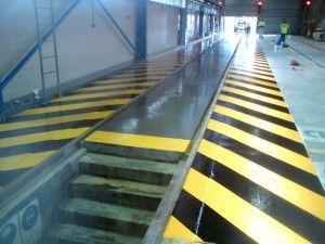 Indoor industrial walkway with black-yellow hazard stripes beside a trench-like pit and stairs