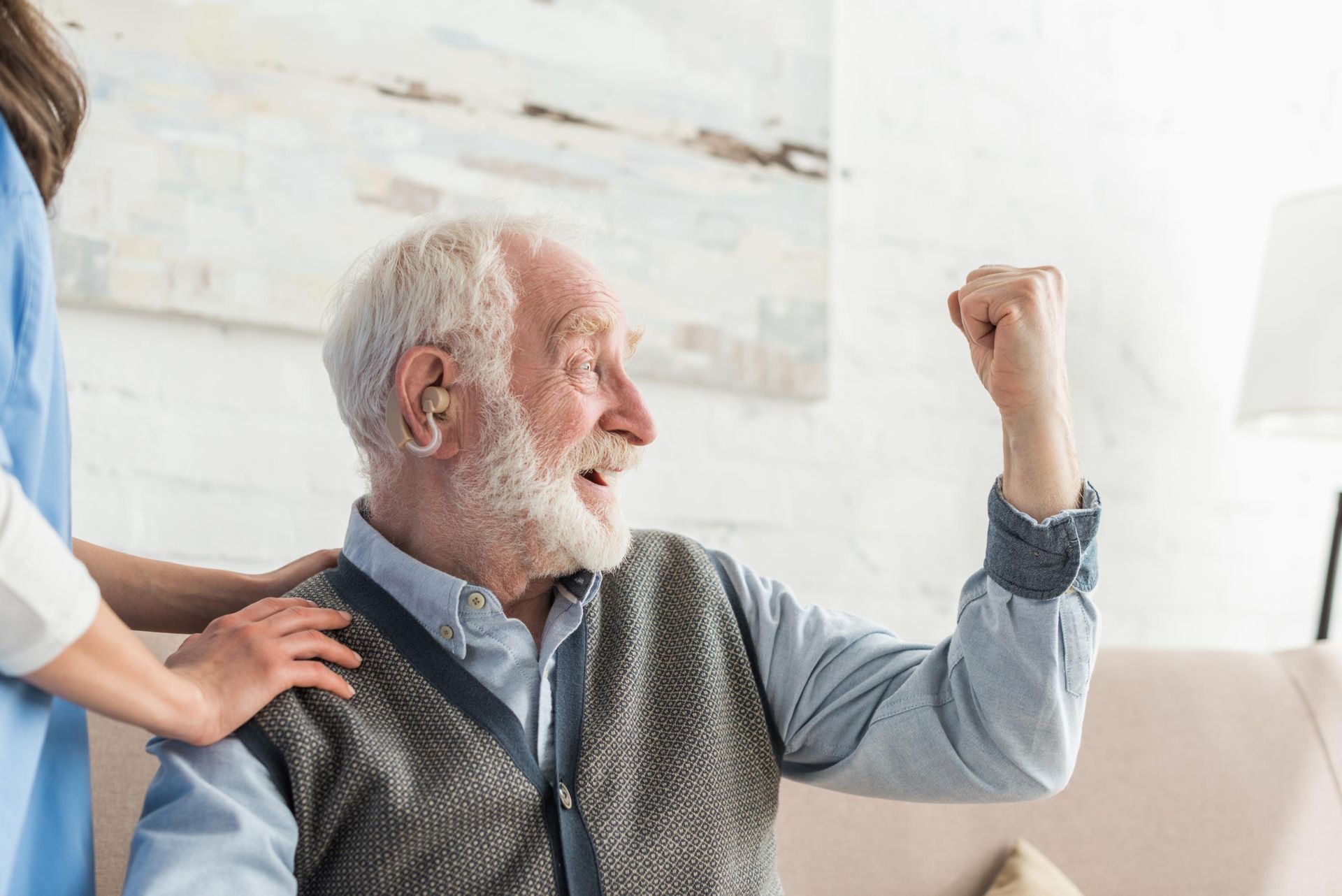Elderly man smiling and raising a fist while a caregiver rests a hand on his shoulder in a bright room