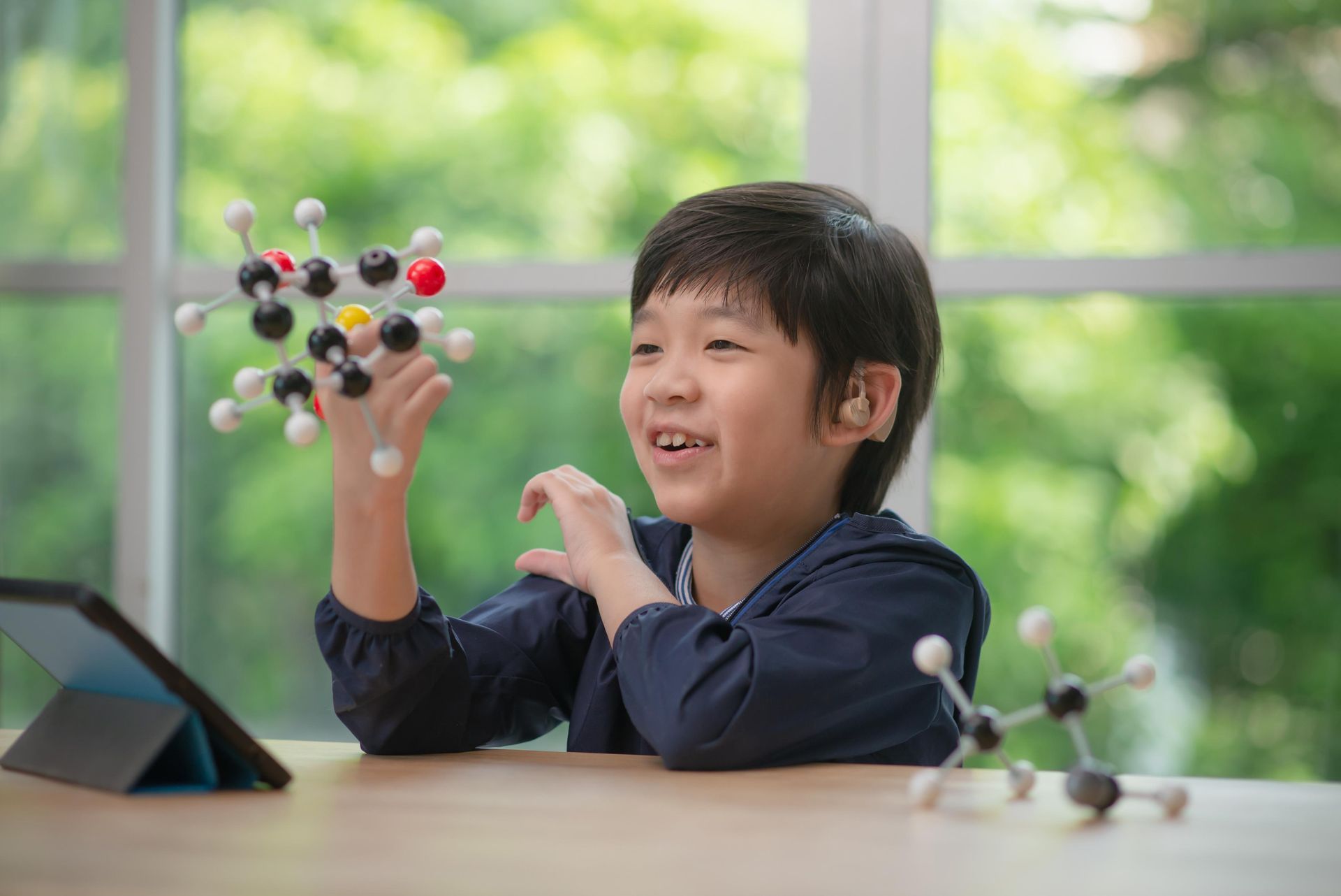 Child examining a molecular model at a table near a window, with a tablet beside them.