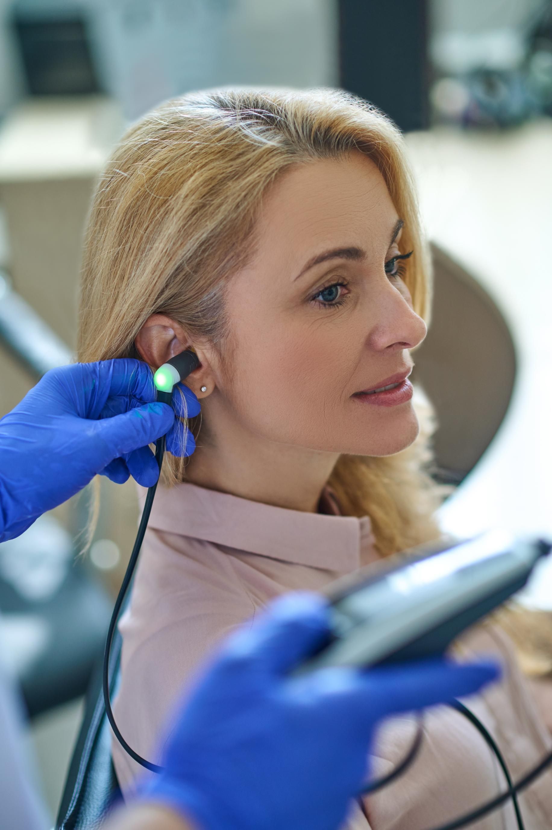 Healthcare worker performing an ear examination on a seated patient with an otoscope and blue gloves