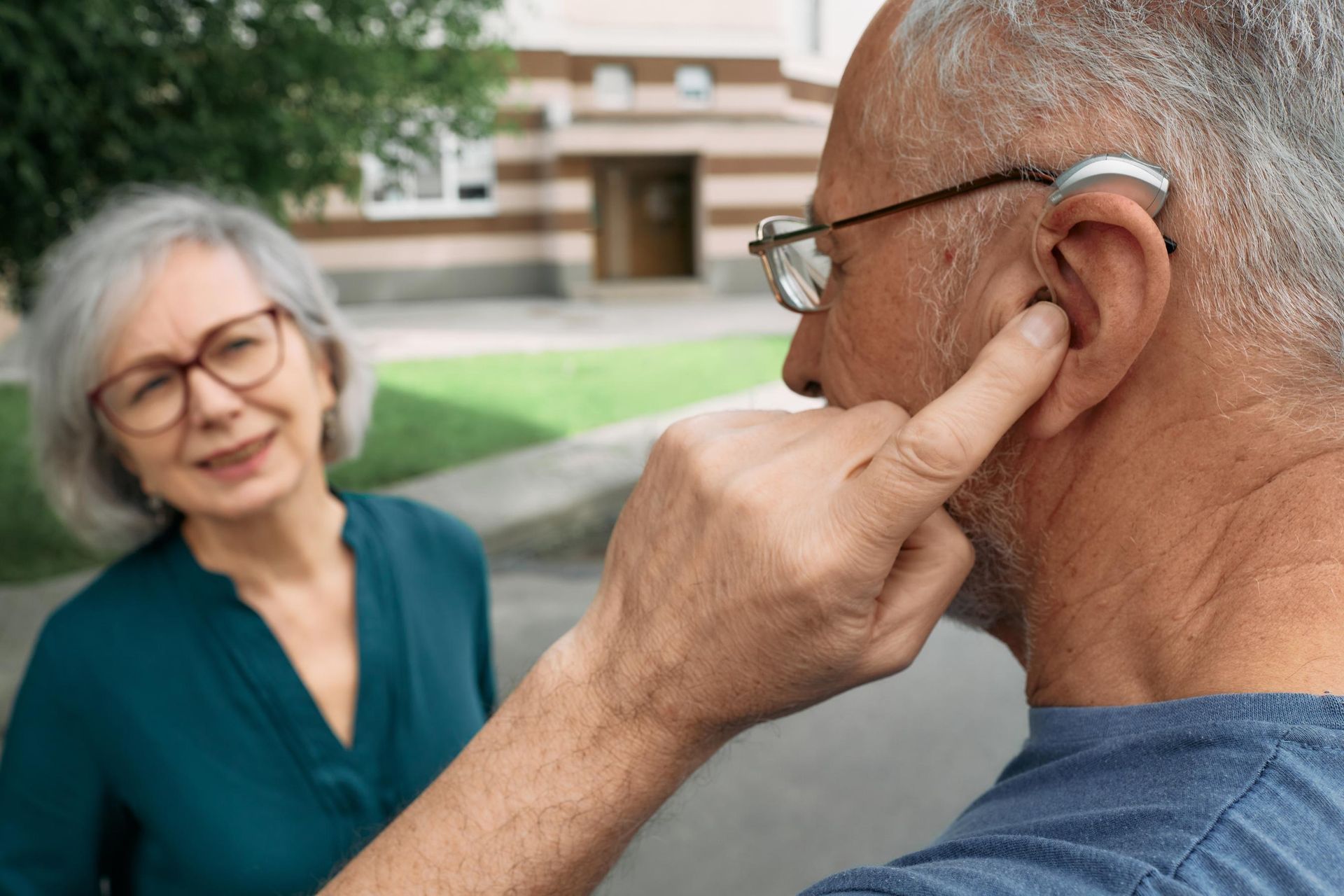 Woman speaking with an older man outdoors, touching his ear near a building.