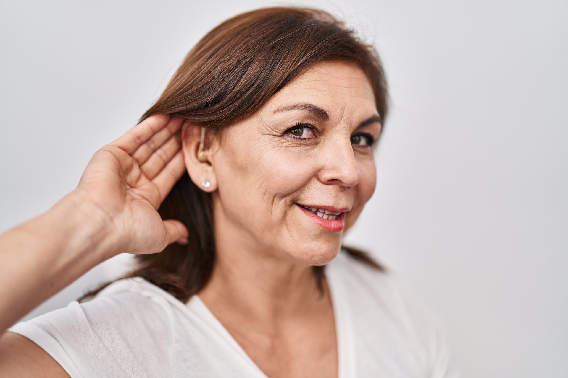 Woman in a white shirt touching her ear and smiling against a light background