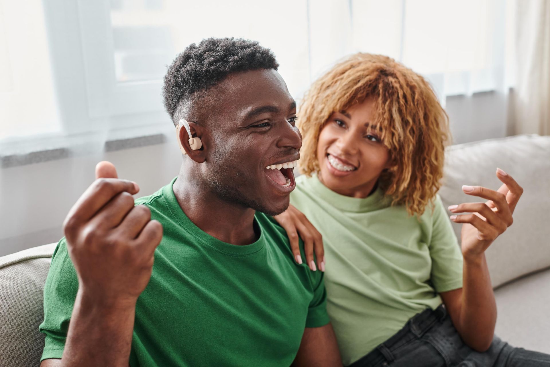 Two people smiling on a couch, one in a green shirt, sharing a warm moment indoors.
