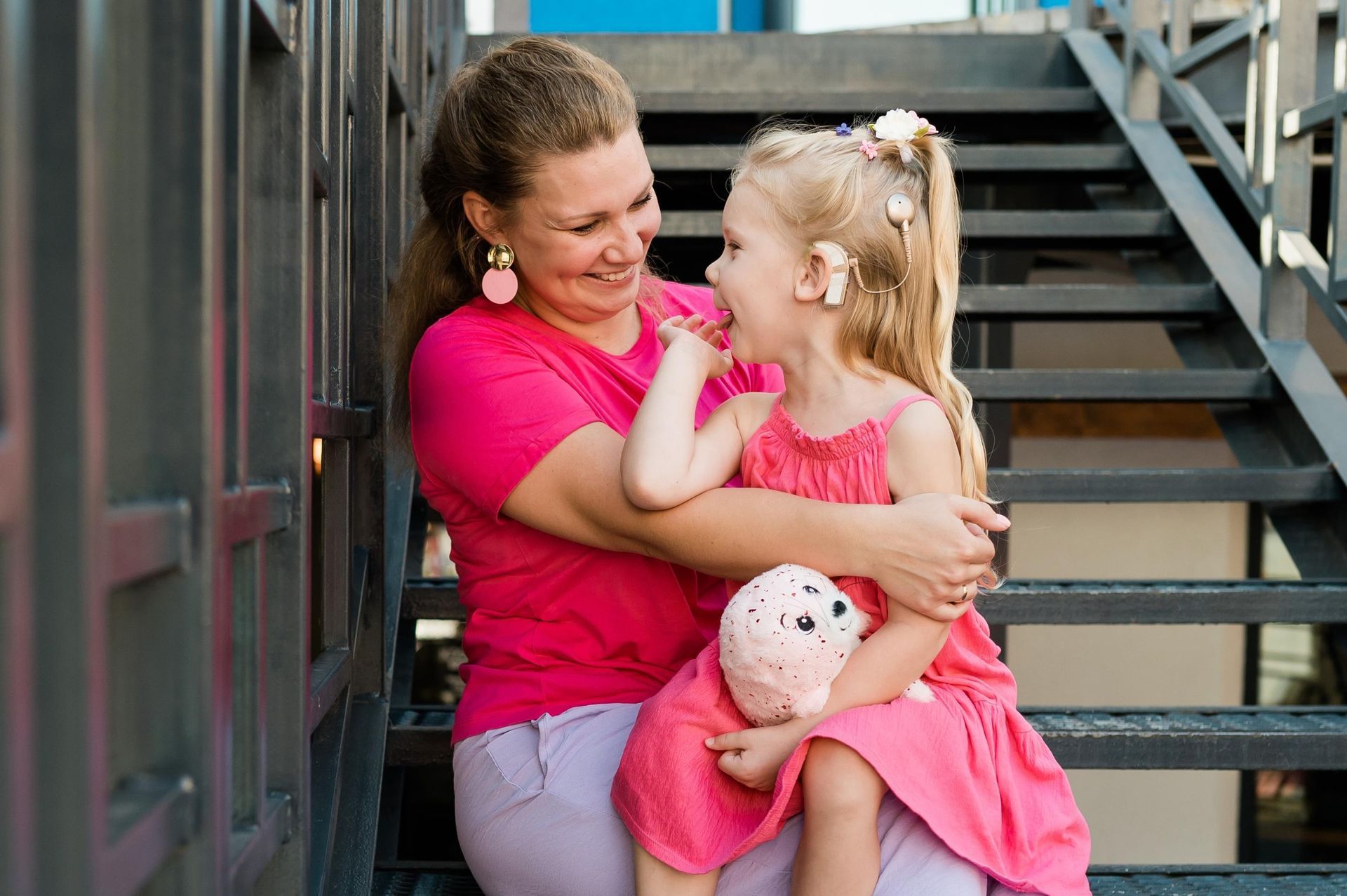 Woman in pink shirt smiling and hugging a child in pink dress on outdoor stairs