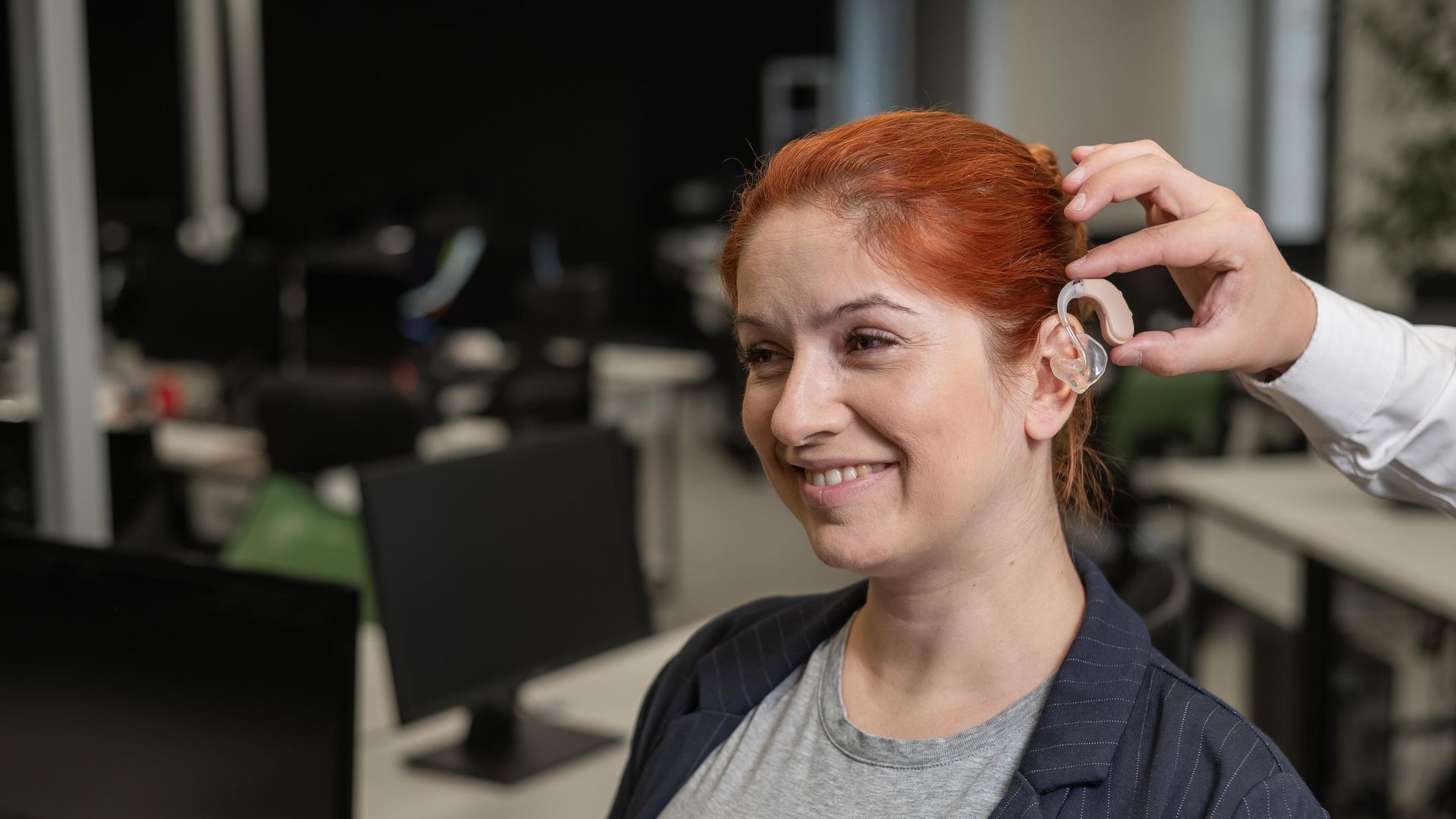 Woman smiling as a hand adjusts a hearing aid in a salon setting