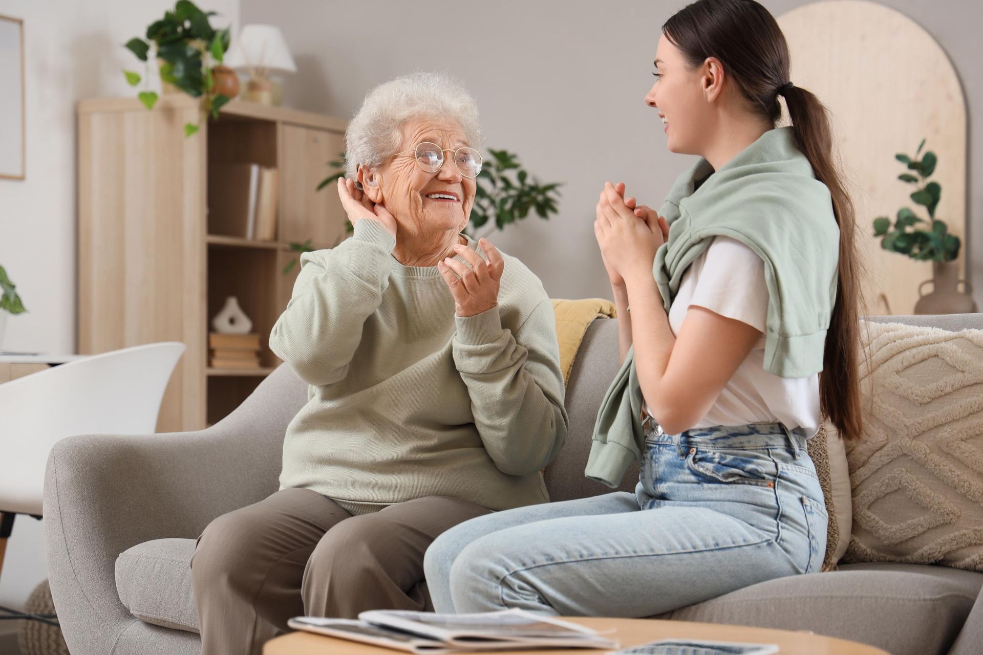 Two people chatting on a sofa in a bright living room, one smiling and gesturing with clasped hands.