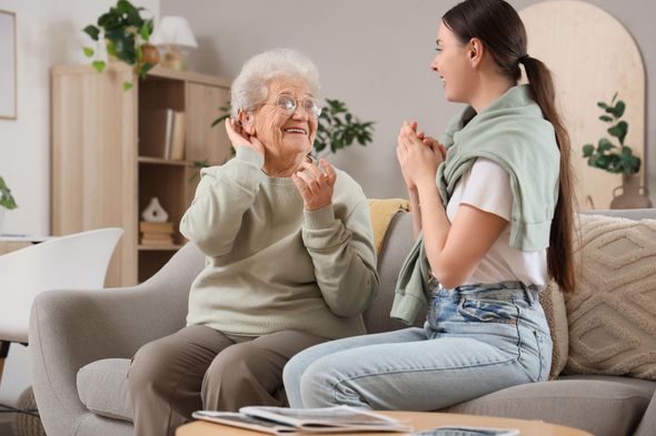 Two women talking on a sofa in a bright living room, one smiling and gesturing.