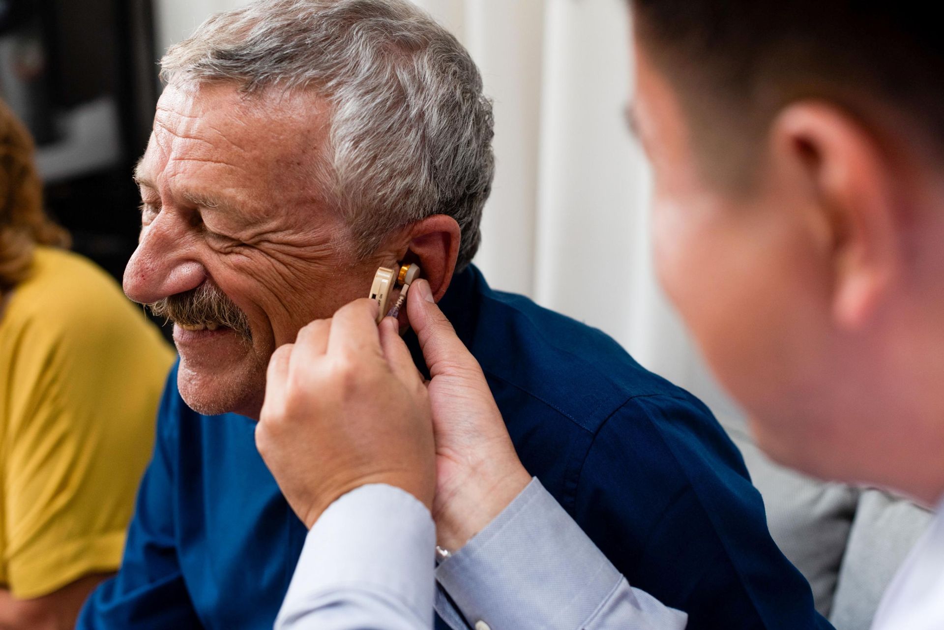 Man fitting a hearing aid for an older man while they sit indoors and talk