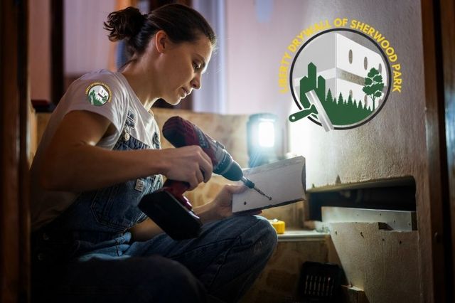Drywall patch repair scene showing a woman kneeling in a dim workspace, holding a red power drill as she secures a small drywall piece illuminated by a work light. She is focused on the repair with tools around her. The residential project is near Sherwood Park, Edmonton, Alberta.