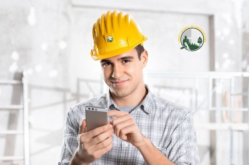 An employee from Liberty Drywall of Sherwood Park wearing a yellow hard hat and plaid shirt holds a smartphone as if checking job details. He stands inside a bright unfinished room, representing drywall work and site coordination in Sherwood Park, AB.