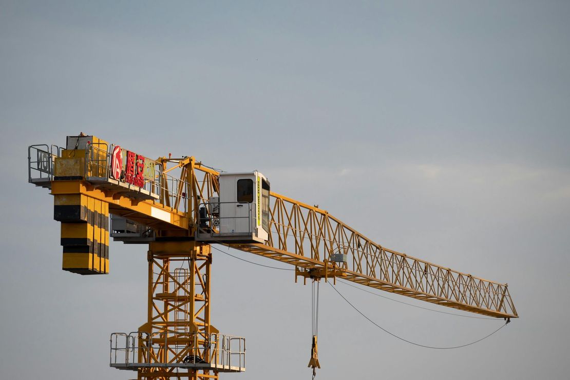 Yellow construction crane against a gray sky, with cables and a suspended hook.